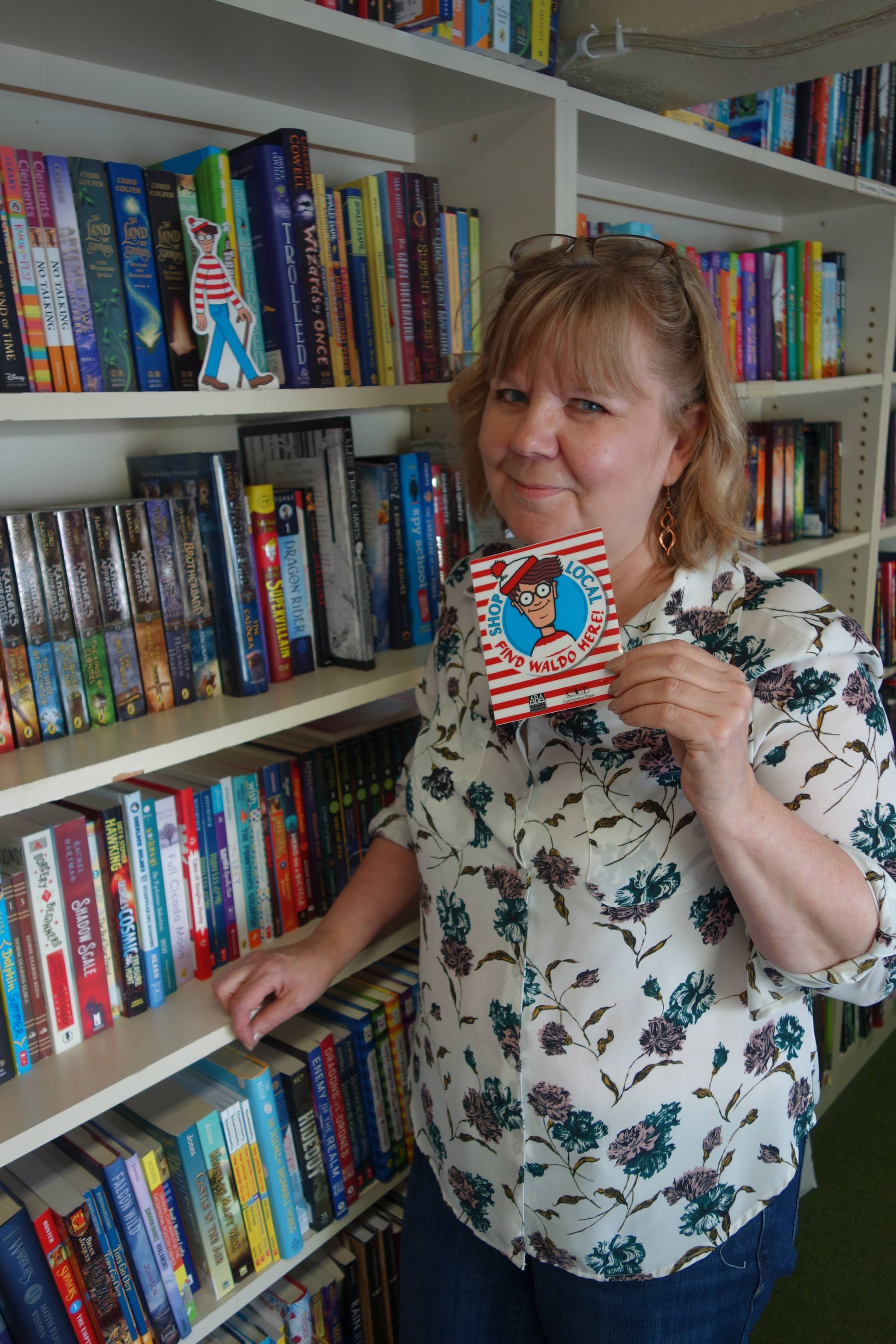 Hearthside Books owner Brenda Weaver holds the Wheres Waldo? sign which will be displayed in downtown Juneau businesses in July to signal to seekers that a six-inch, cardboard Waldo will be hiding in the store. In this July 21 photo, Waldo can be seen standing on the bookshelf. Clara Miller | Capital City Weekly