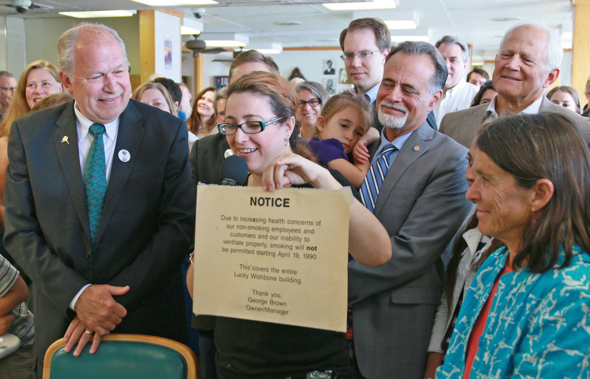 In a photo provided by the office of the governor, Gov. Bill Walker signs Senate Bill 63, a statewide public smoking ban, on Tuesday, July 17, 2018 at the Lucky Wishbone restaurant in Anchorage. Standing at right is Sen. Peter Micciche, R-Soldotna, the legislative sponsor of the bill. On Walker’s lap is Stella Micciche, one of Micciche’s daughters. Also visible, from right to left behind Walker are Dr. Jay Butler, Alaska’s chief medical officer; Rep. Geran Tarr, D-Anchorage; Rep. Jason Grenn, I-Anchorage; Micciche; Rep. Chris Birch, R-Anchorage; and the smile of Emily Nenon of the American Cancer Society. (Courtesy photo)
