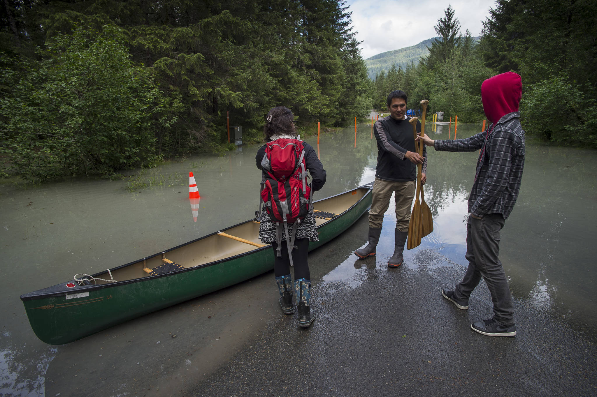 Anthony Mallott, center, hands out paddles as he brings a canoe to help people across a flooded section of View Drive on Thursday, July 19, 2018, after a release of water from Suicide Basin on Wednesday. (Michael Penn | Juneau Empire)