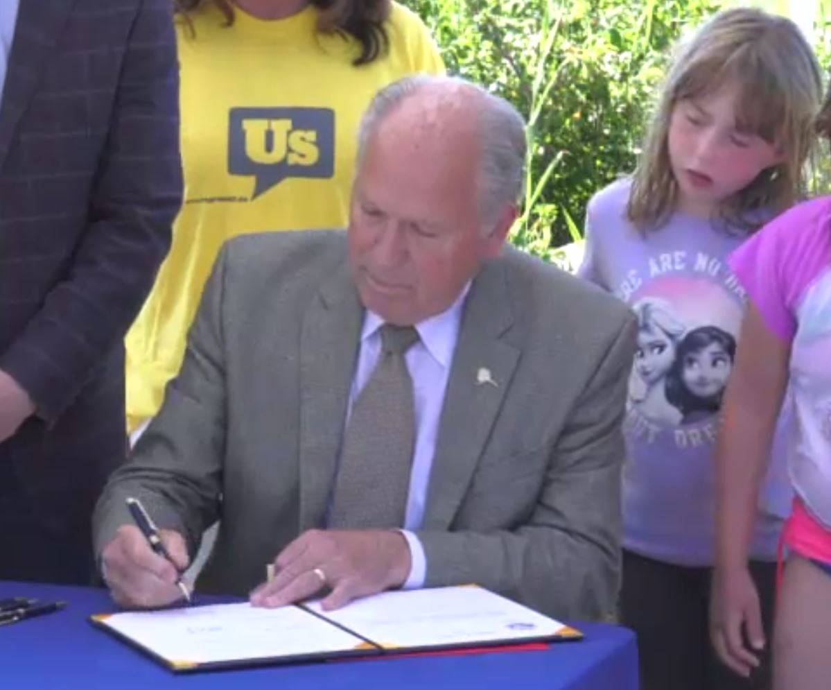 Gov. Bill Walker signs House Bill 44 in a ceremony under a temporary tent Thursday evening at Jewel Lake in Anchorage while a curious child looks on. (Screenshot)