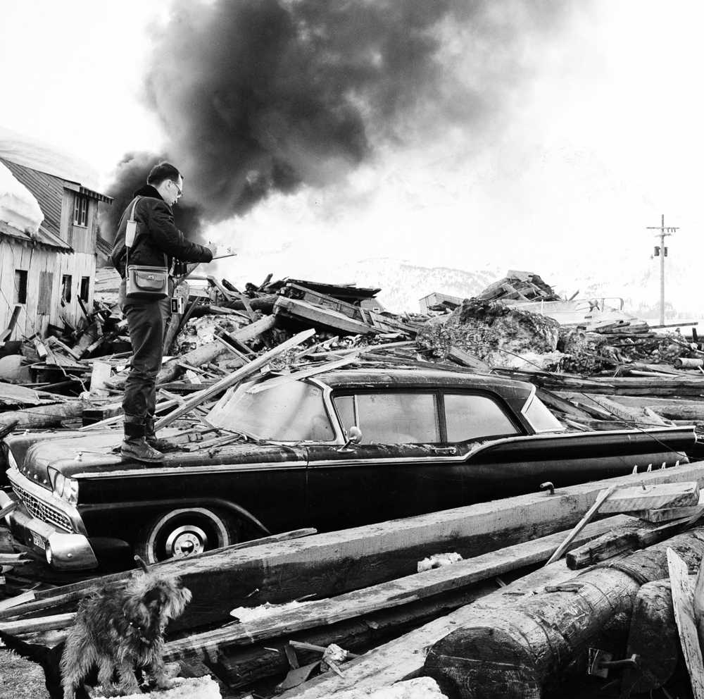 FILE - In this March 29, 1964 file photo, a photographer looks over wreckage as smoke rises in the background from burning oil storage tanks at Valdez, Alaska. On Monday, Feb. 1. 2016, federal scientists say they've pinpointed the cause of tsunami waves following the 1964 Great Alaska Earthquake, the second-largest ever recorded, at magnitude 9.2. (AP Photo/File)