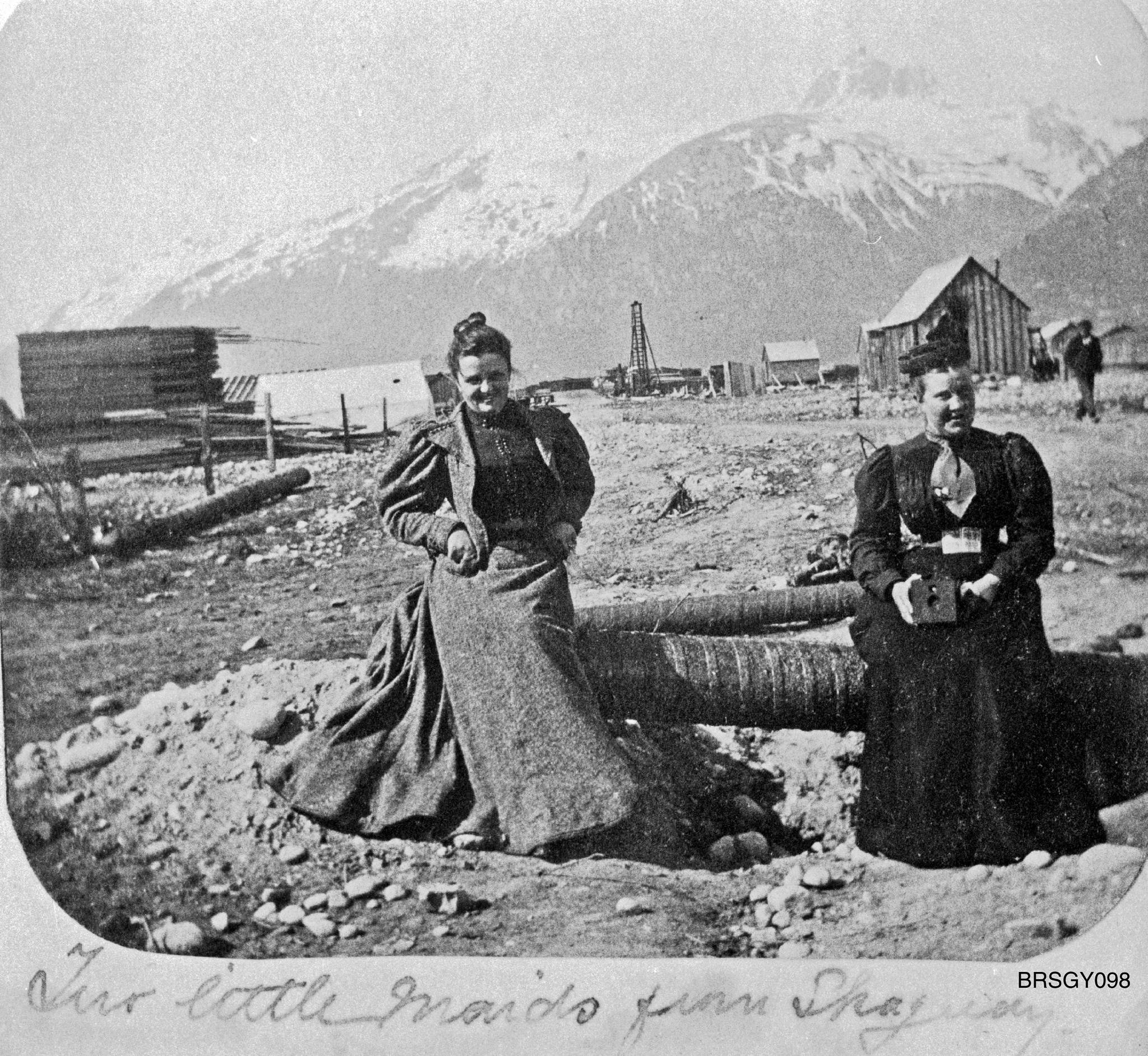 “Two little maids from Skagway” looking southwest down Main Street, circa summer 1898. Mollie Brackett, left, and Mrs. Tuckerman sit on a wood stave pipe, which was about to be installed on Main Street, possibly at the cross street of 3rd Ave. Tuckerman is holding a woodbox camera probably similar to the one Brackett used. (Courtesy Photo | National Park Service, Klondike Gold Rush National Historical Park, Brackett Family Collection, BRSGY089, KLGO SP-143-5368)