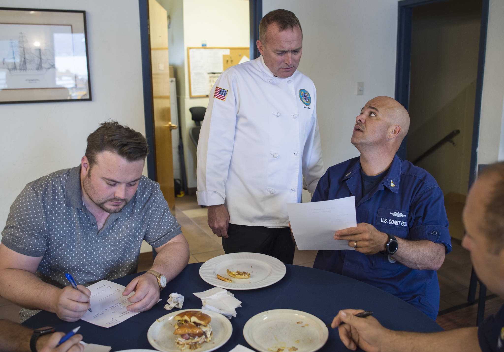 Master Chief Justin Reed, center, answers a question by Senior Chief Jeremy Demello, right, as Jared Curé, owner of The Narrows, left, judge a best burger contest between four teams of U.S. Coast Guard buoy tender chefs on Thursday, Aug. 23, 2018. (Michael Penn | Juneau Empire)