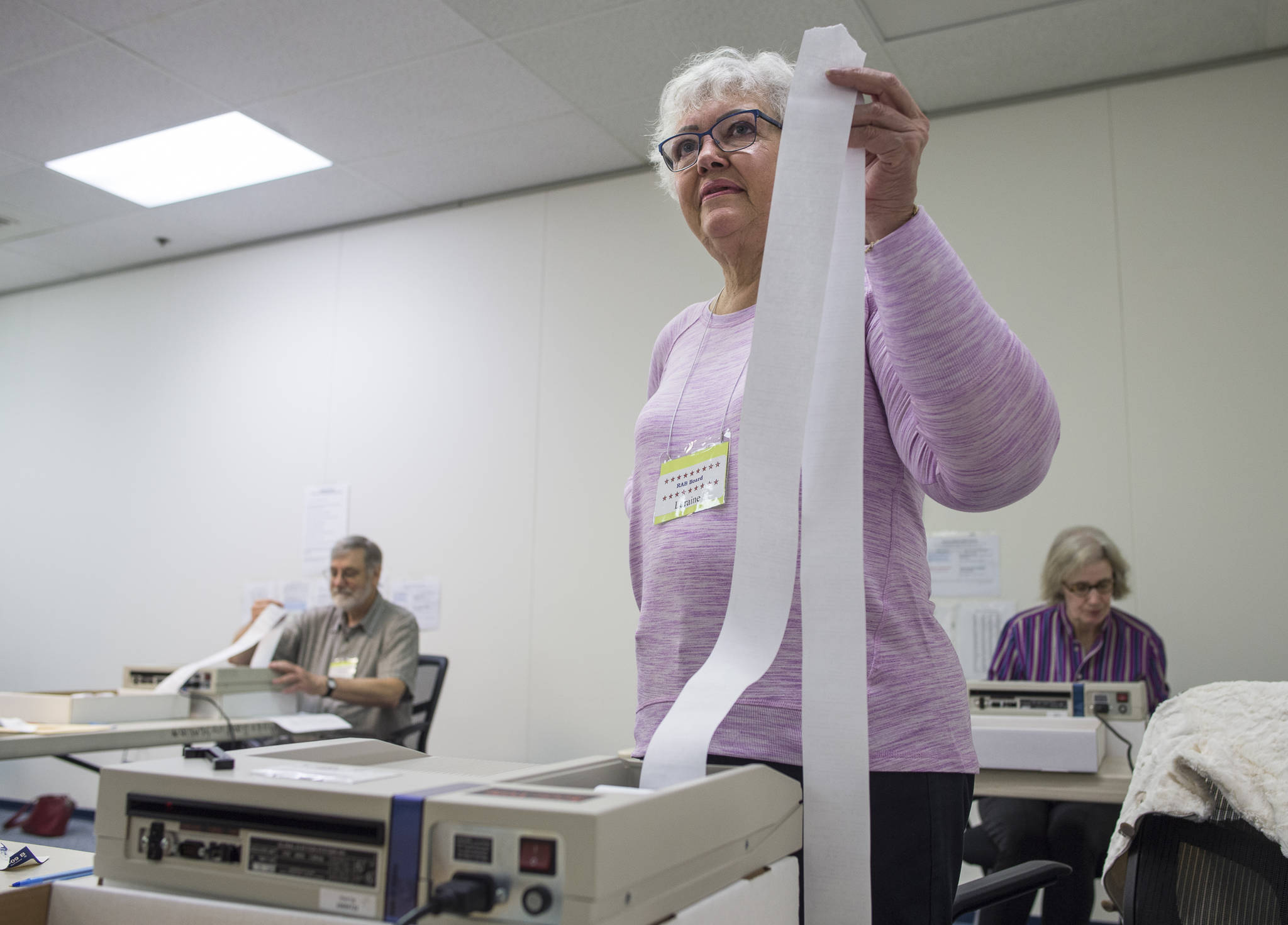 Laraine Derr, of the Regional Accu-Vote Board, center, waits for her machine to print out a tally as she, David Clover, left, and Mary Foster count questioned and absentee ballots at the State of Alaska election office at the Mendenhall Mall on Tuesday, Aug. 28, 2018. (Michael Penn | Juneau Empire)