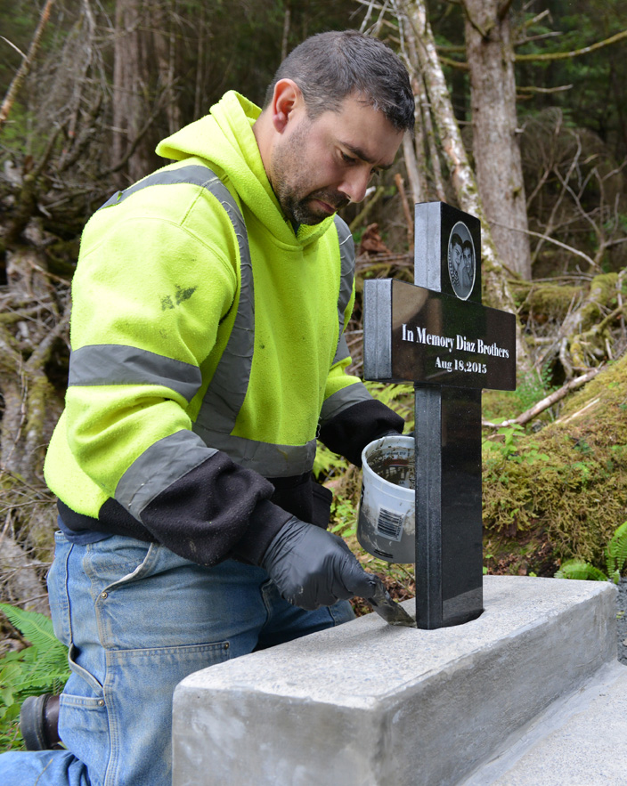 JR Lindstrom, a close friend of the Diaz brothers, cements a cross – with the engraved photos of Elmer and Ulises – in place near a memorial bench Wednesday evening. More than 200 people gathered for the dedication of  the bench and cross Thursday afternoon at the head of the Cross Trial near Kramer Avenue. The bench, which affords views over Sitka Sound and Mt. Edgecumbe, was dedicated to the memory of Elmer and Ulises Diaz and William Stortz who died in the Aug. 18, 2015, landslide on Kramer Avenue.