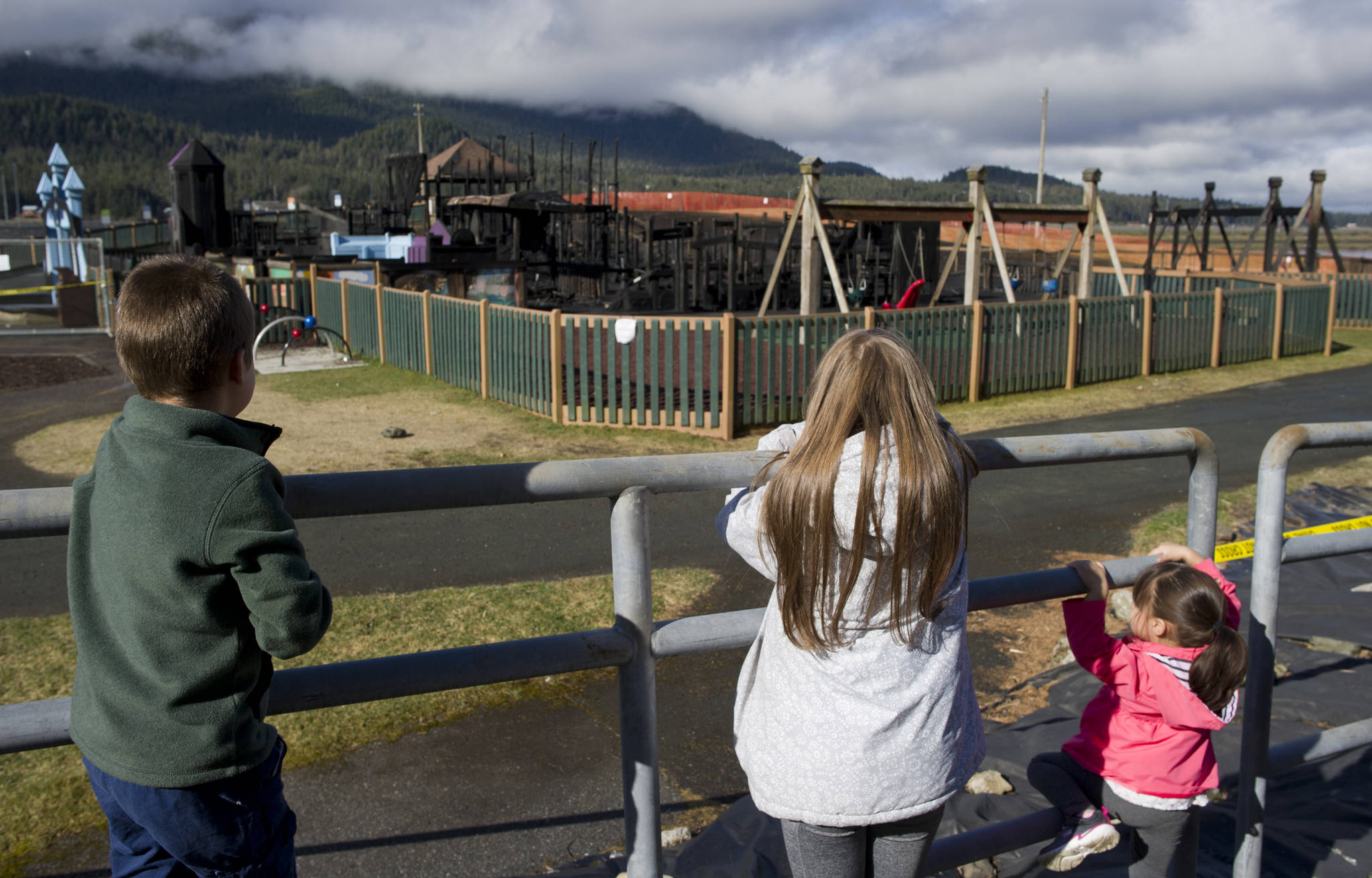 Sibblings Marco Carranza, 5, left, Monica, 11, center, and Kaitlyn, 3, view Project Playground from behind police tape on Tuesday. A fire set by two 13-year-old boys destroyed the playground Monday. (Michael Penn | Juneau Empire)