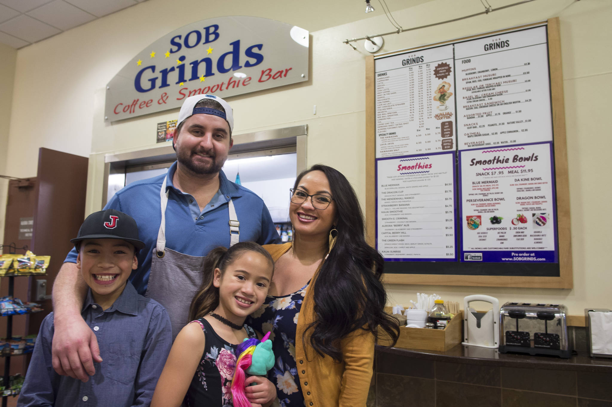 Erik Scholl with his partner, Tianna Banua, and their children Ezra and Ezlyn Vidal, stand in front of one of their two food businesses in the State Office Building on Thursday, Nov. 9, 2017. They operate SOB Grinds and Shaka Shack, both located on either end of the eighth floor atrium. (Michael Penn | Juneau Empire)
