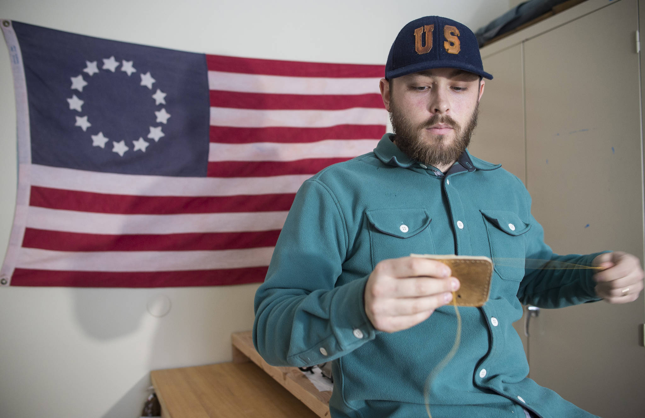 In front of a replica 1776 U.S. flag, KC Mack hand sews a wallet from the leather of a used baseball mitt on Friday, Nov. 17, 2017. (Michael Penn | Juneau Empire)