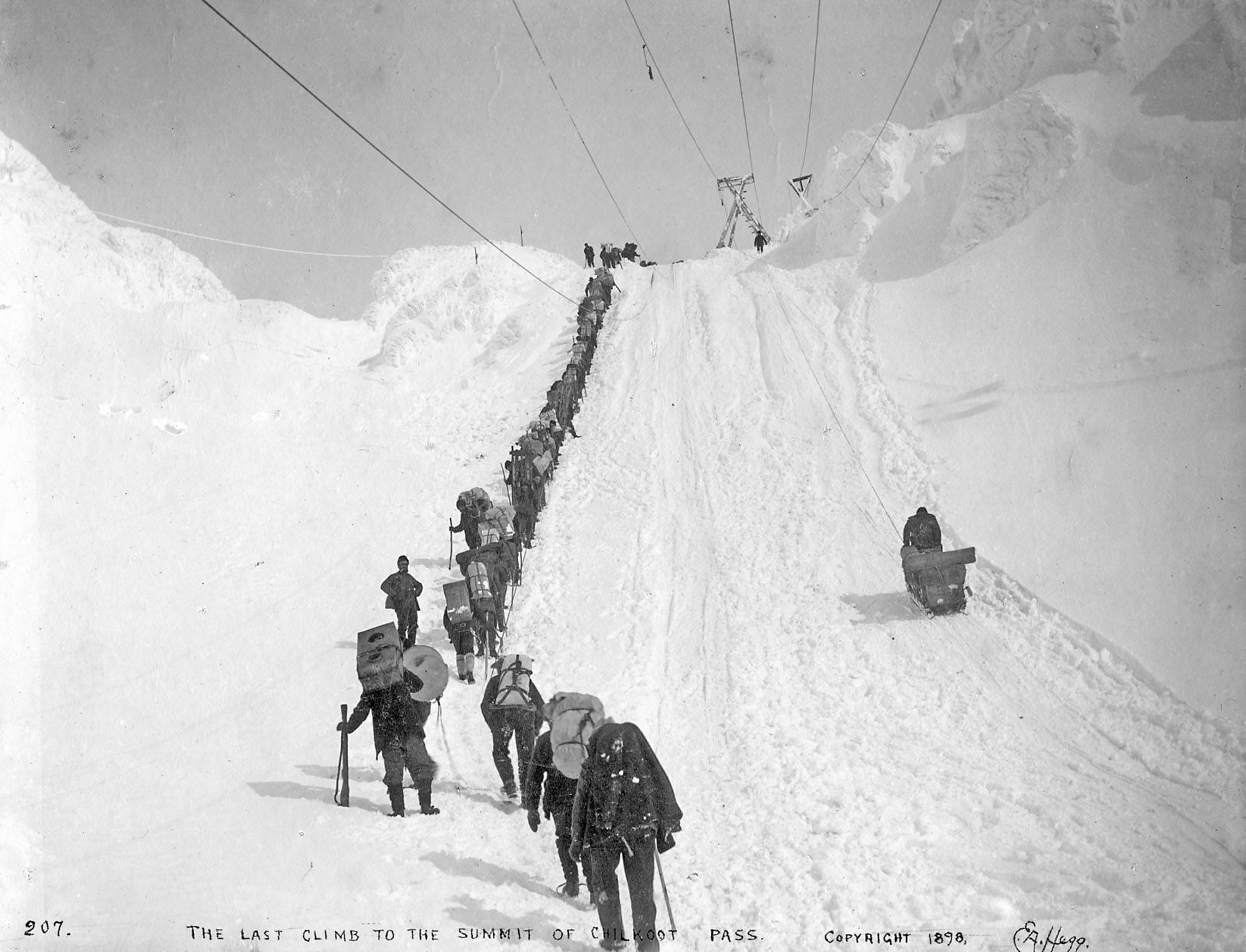 National Park Service, Klondike Gold Rush National Historical Library, Darcie Culbeck Collection, KLGO CS-50-10573. “The Last Climb to the Summit of Chilkoot Pass,” taken winter 1898. Photographer: Eric A. Hegg (207). In the center left is the northern end of the Golden Stairs between the False Summit and the True Summit and to the right of that is a single sled probably being hauled up to the Summit by the gasoline winch, part of Archie Burns’ surface tramway system. The gasoline-powered tramway was introduced by mid-April 1898. It was described as “simply a pulley drum and gasoline engine at the summit of the pass, and enough rope to reach the bottom. Sleds were hitched onto the rope, which was wound around the drum and it pulled them to the top.”