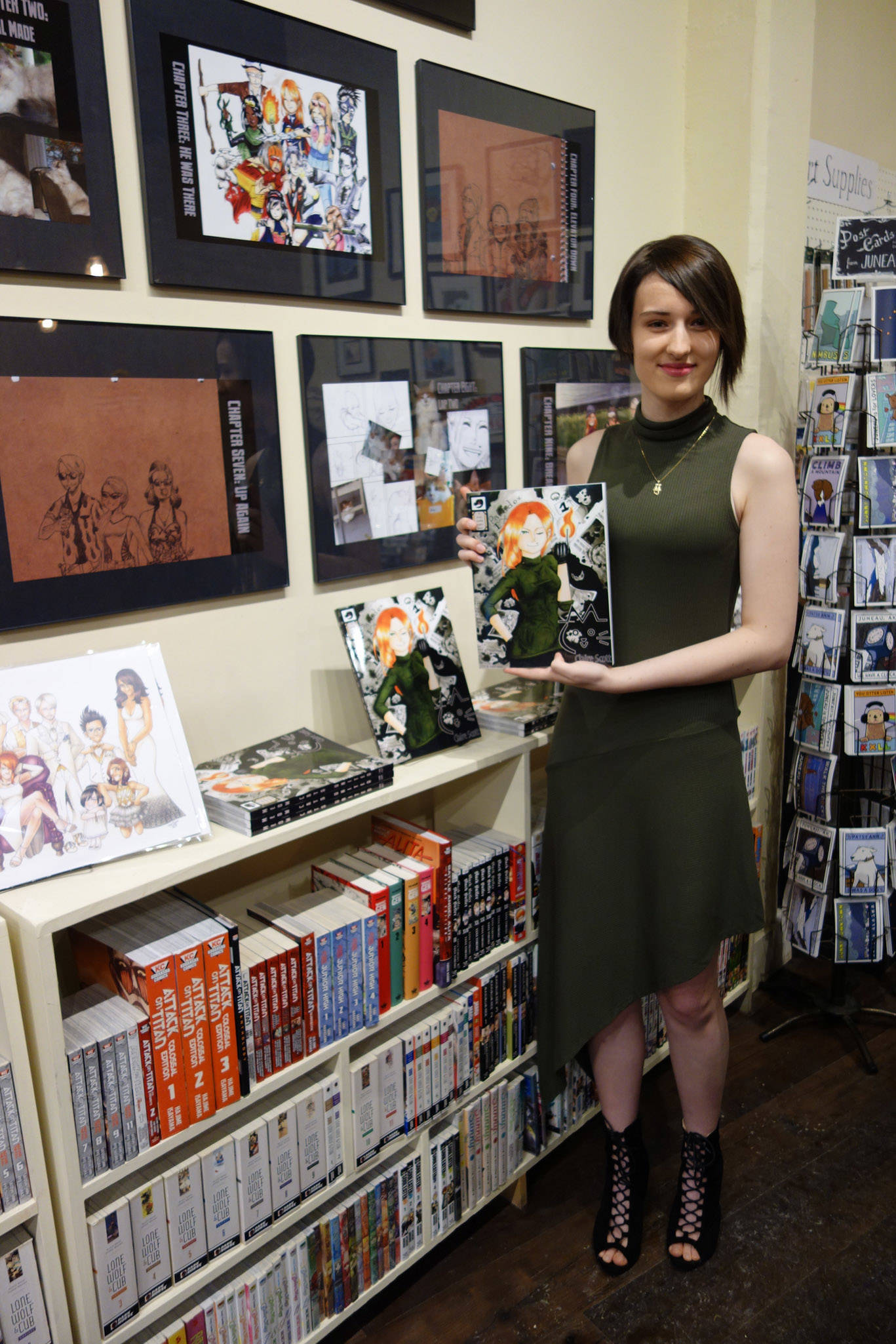 Claire Scott poses with a copy of her graphic novel “Meow Cats United” in front of her art on display in the Alaska Robotics Gallery on First Friday. Clara Miller | Capital City Weekly