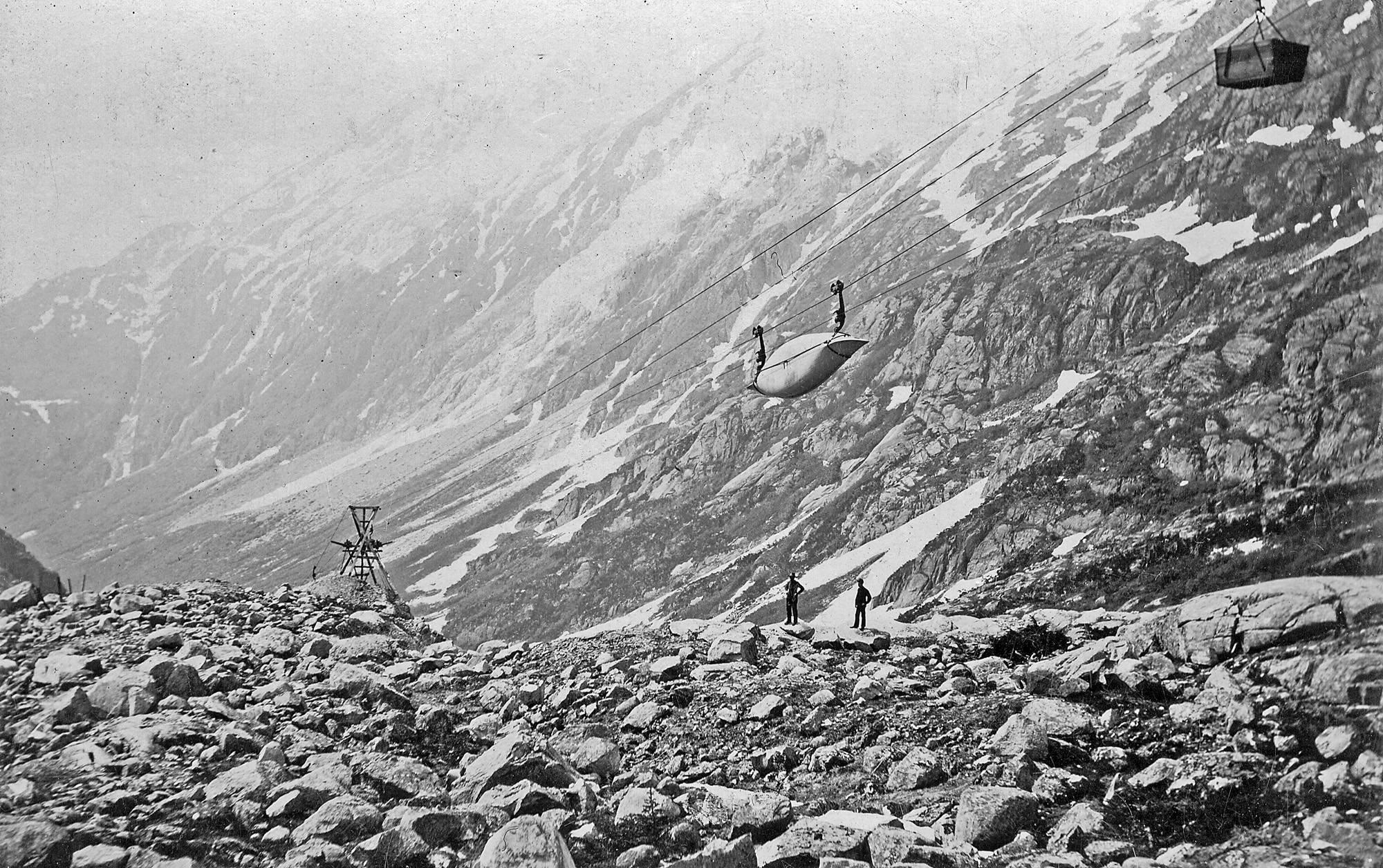 Long Hill, Chilkoot Trail, looking southwest. This view shows the Chilkoot Railroad & Transport Company (CR&T) aerial tramway in operation. One of the CR&T tramway towers is in the background left and a canoe and crate are being hauled over the line toward the summit of the Chilkoot Trail. Two men, possibly tramway workers, are observing the load. This photograph was taken on Long Hill between Sheep Camp and the Scales between the spring-fall of 1898-1899. Image courtesy of the National Park Service, Klondike Gold Rush National Historical Park, Candy Waugaman Collection, KLGO LH-68-8973.