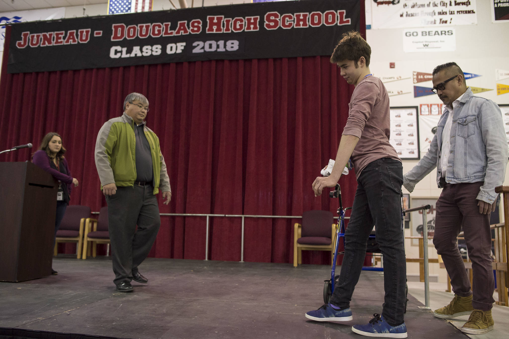Henry Cheng, a graduating senior at Juneau-Douglas High School, practices walking across the stage at JDHS with the help of his father, Vincent, second from left, para educator Valerie Cables, right, and physical therapist Che Plang, of Southeast Alaska Therapies, on Thursday, May 24, 2018, in preperation of Sunday’s graduation ceremony. Henry suffered a severe traumatic brain injury after falling off a cliff in 2016. (Michael Penn | Juneau Empire)