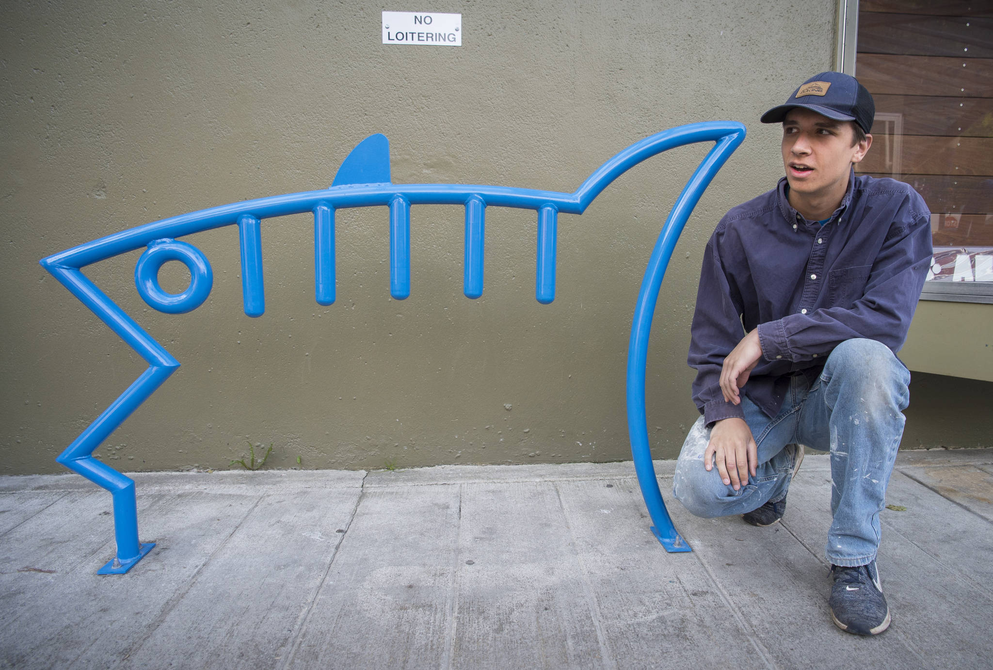 Ben Campbell, a recent graduate of Juneau-Douglas High School, kneels next to one of two of his fish designed bicycle racks on Ferry Way on Wednesday, June 13, 2018. Campbell designed the rack for a class and the city choose to make it a reality. (Michael Penn | Juneau Empire)