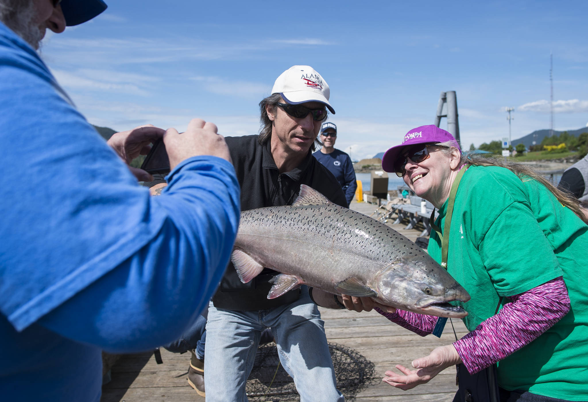 Paula Varney, right, has her picture taken by her husband, Brad, with an estimated 25-pound king salmon held by fishing guide Paul Turinsky, of Chum Fun!, at the Wayside Park on Channel Drive on Wednesday, June 13, 2018. The fish was released after the picture but king salmon can be kept starting today. (Michael Penn | Juneau Empire)