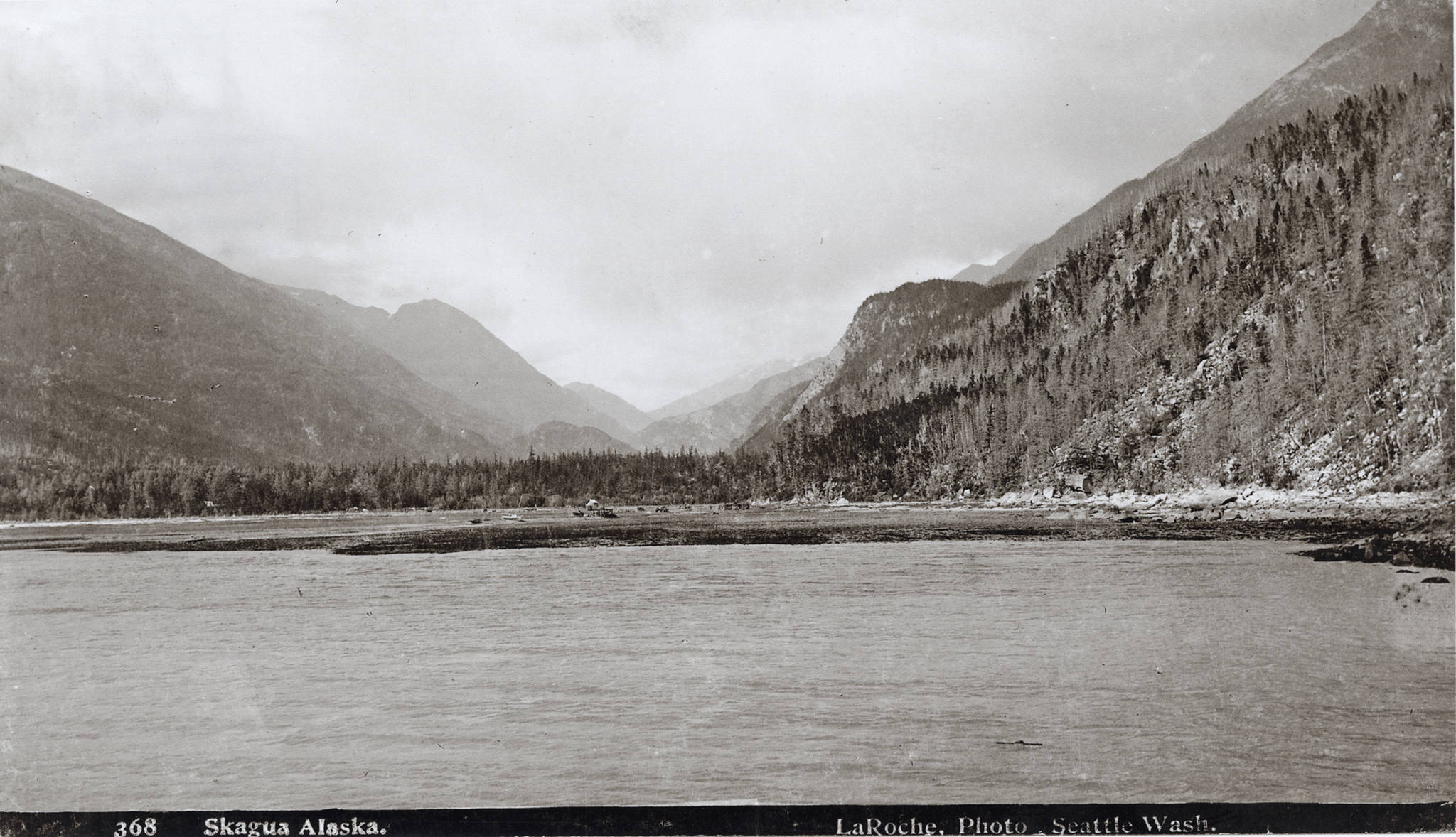 This photograph of the Skagway valley was taken from the deck of the Steamer Queen on July 26, 1897 and shows the almost undisturbed terrain of the Skagway Valley just before the Klondike Gold Rush begins for Skagway. This is Mooresville, Captain Moore’s name for his little hamlet. In the next few hours, days, weeks, and months, the gold seeking Argonauts will invade the valley like an army with their outfits, tents and animals. In less than a month, a street grid will be laid out. The tents of the first month will quickly give way to wood buildings, one, two, and three stories high and the population will increase from around two dozen or so to over 10,000 by the start of 1898. In the center of this image stand Captain William Moore’s Cabin from 1887 and in front, his son Ben Moore’s wood-frame house under construction. (Library of Congress, 3c22304u; KLGO SE-9-8799)