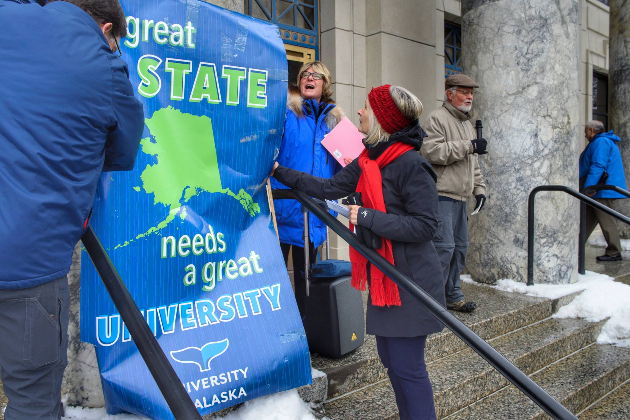University of Alaska Southeast Instructor Jim Powell, left, his wife, former Rep. Beth Kerttula, and Rep. Andi Story, D-Juneau, right, stand up a university sign as former Chancellor John Pugh speaks at a rally for funding the University of Alaska in front of the Capitol on Wednesday, Feb. 13, 2019. (Michael Penn | Juneau Empire)