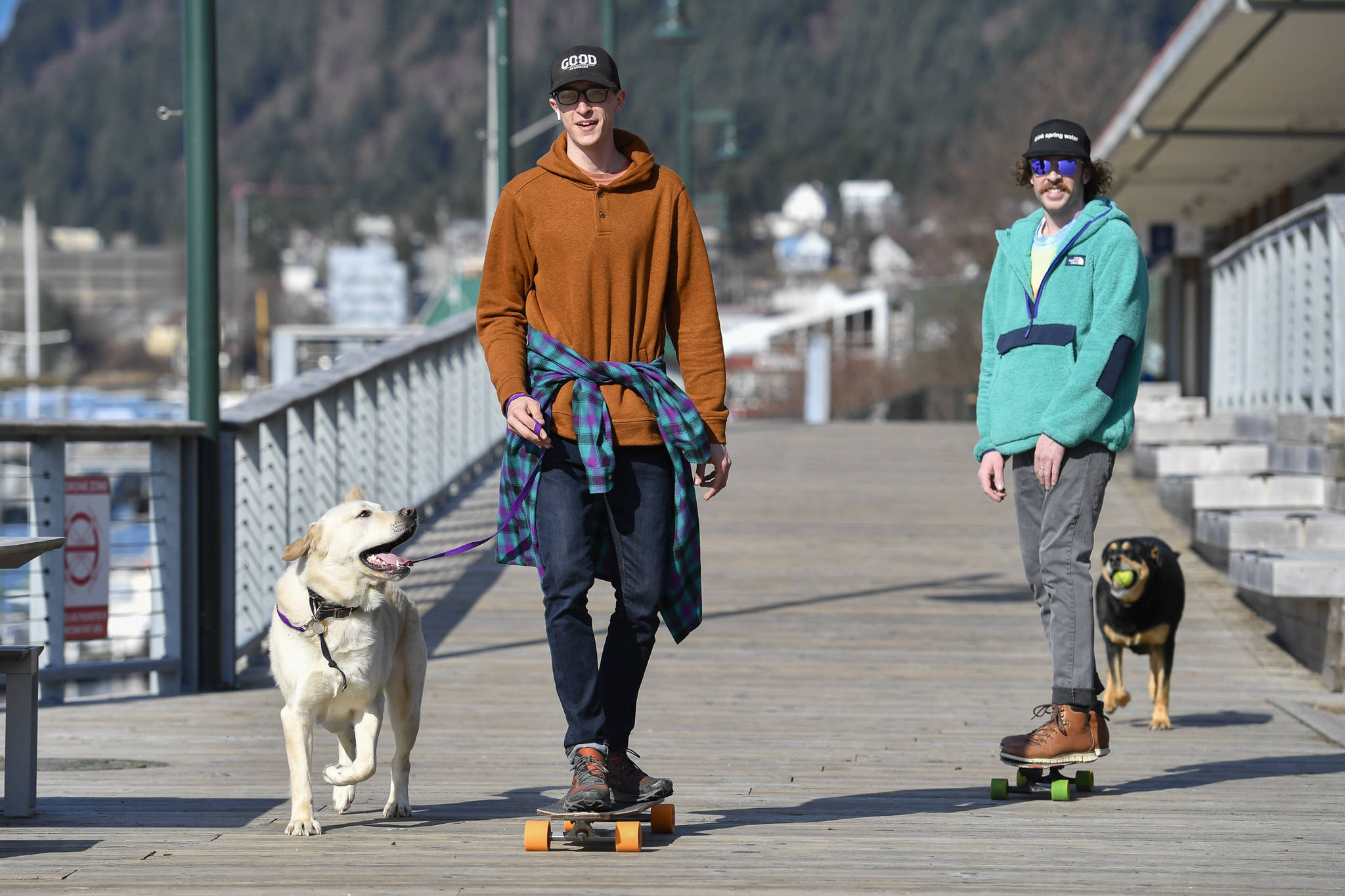 Cody Kopkie, left, with Olly, and Ray Huebschen, followed by Nala, take advantage of the sunny weather and lack of ice to skateboard along the Seawalk on Monday, March 25, 2019. (Michael Penn | Juneau Empire)