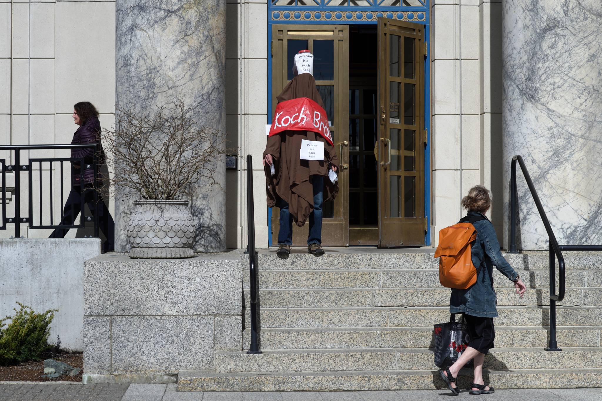 An anonymous protester, dressed as a Coke bottle, hands out moose poop as Gov. Mike Dunleavy ‘s budget to Sue Christiansen on the steps of the Capitol on Monday, April 15, 2019. The protester said, “Oh you’ve got to have a little humor in the whole thing because it’s starting to be a sad country. People don’t make laws anymore, it’s corporations that do for their interests.” (Michael Penn | Juneau Empire)