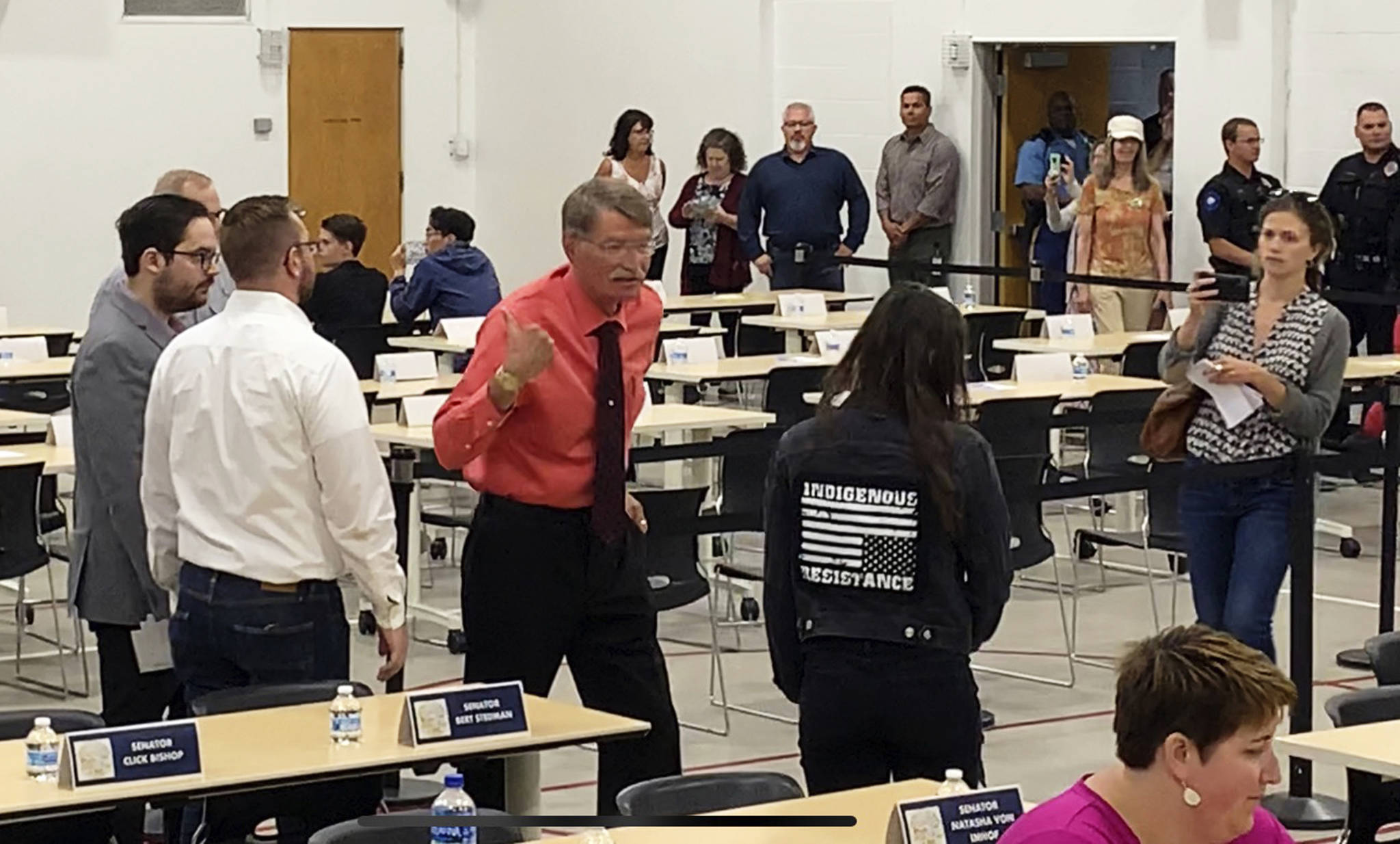 In this July 10, 2019, photo provided by Rochelle Adams, the mayor of Wasilla, Alaska, Bert Cottle, center left in the orange shirt, is seen after grabbing the arm of Alaska Native activist Haliehana Stepetin, who was trying to take part in a sit-in of legislators meeting in Wasilla. Stepetin filed an assault complaint with the Wasilla Police Department on Thursday, naming Cottle and Zachary Freeman, the spokesman for the Republican House Minority, seen next to Cottle in the white dress shirt. (Rochelle Adams via AP)