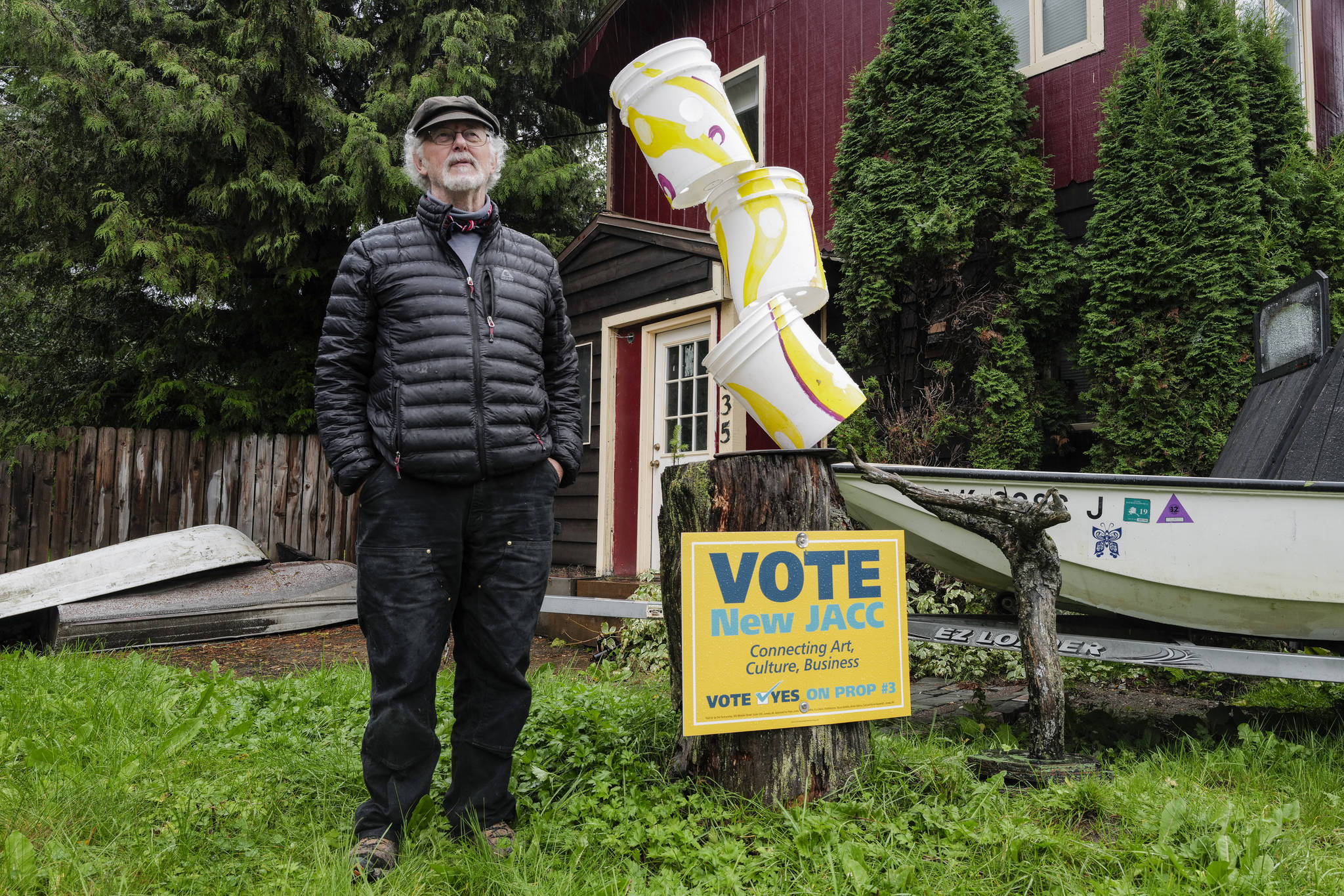 Juneau artist Arnie Weimer stands next to his bucket sculpture and a pro-JACC sign in front of his 12th Street home on Friday, Sept. 20, 2019. (Michael Penn | Juneau Empire)