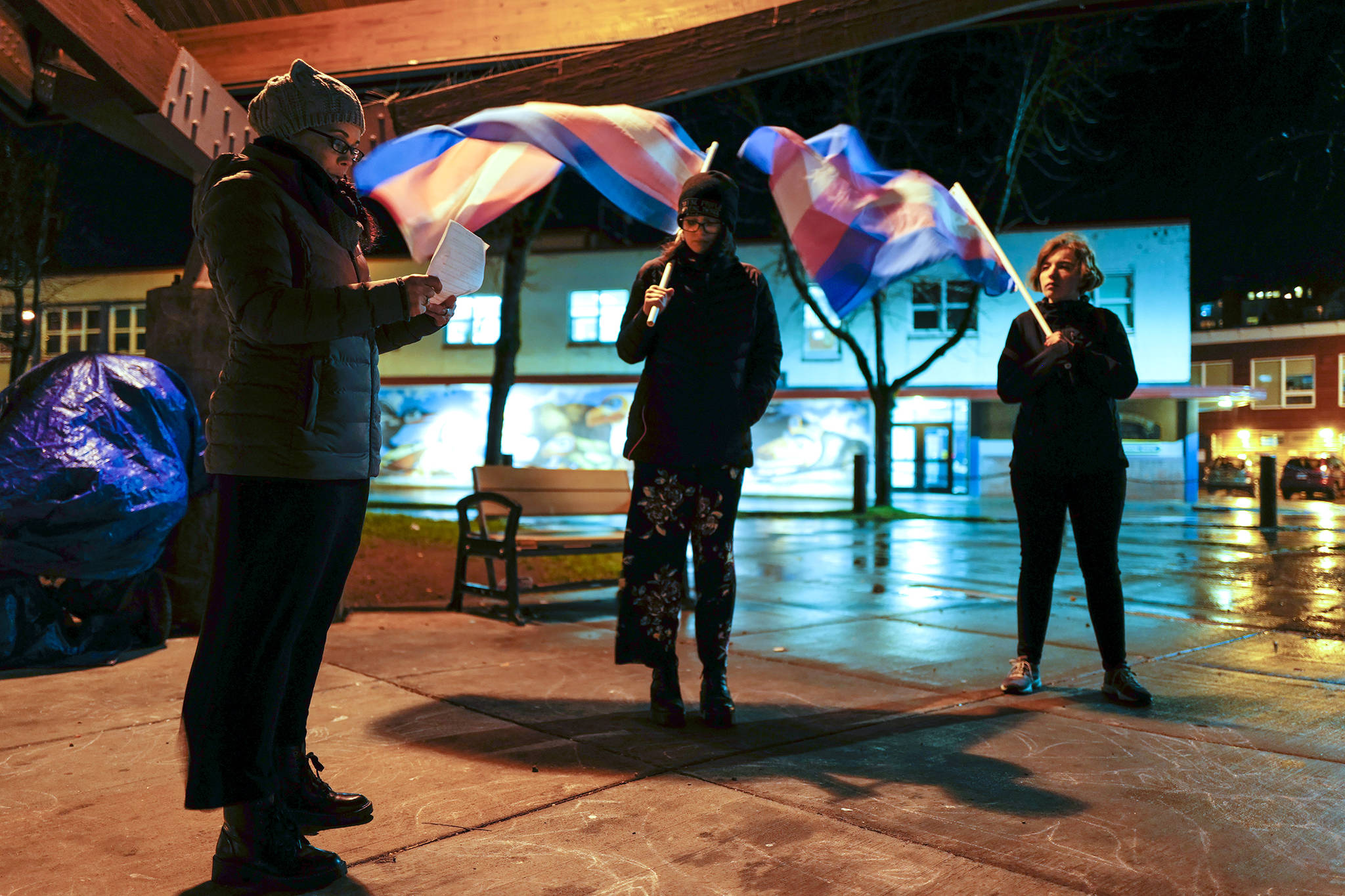 Southeast Alaska LBTQ+ Alliance Board Chair JoLynn Shriber reads a list the names of killed transgender people as Thunder Mountain High School students Kyla Stevens, center, and Laila Williams hold flags in the wind during a transgender remembrance at Marine Park on Wednesday, Nov. 20, 2019. (Michael Penn | Juneau Empire)