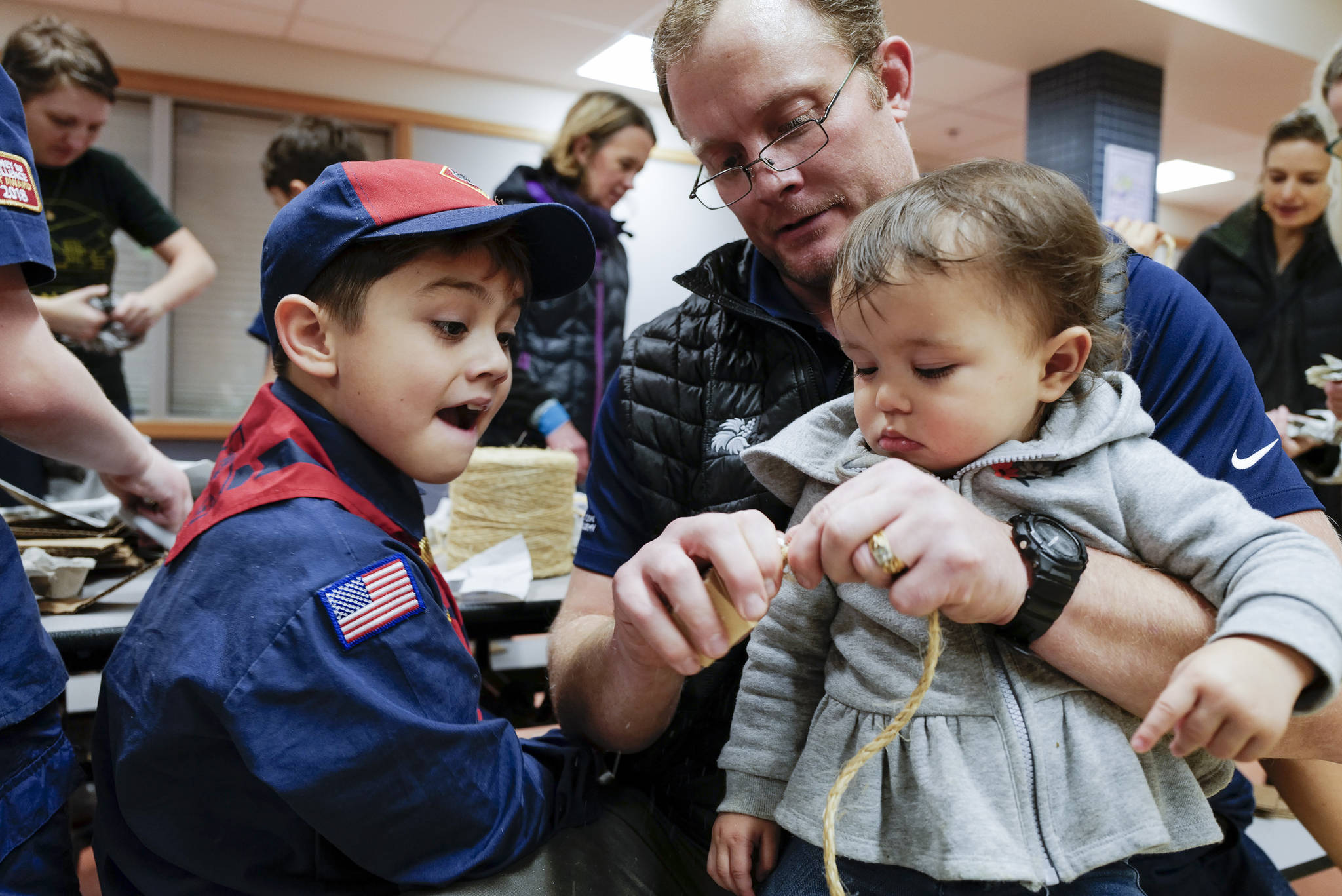 Michael LaRue helps his son, Joseph, 8, left, with his enrichment toys for birds while holding his 15-month-old daughter, Mylene, during a Cub Scout Pack 10 meeting at Harborview Elementary School on Friday, Nov. 22, 2019. About 25 cub scouts constructed the toys as a service project for the Juneau Raptor Center. (Michael Penn | Juneau Empire)