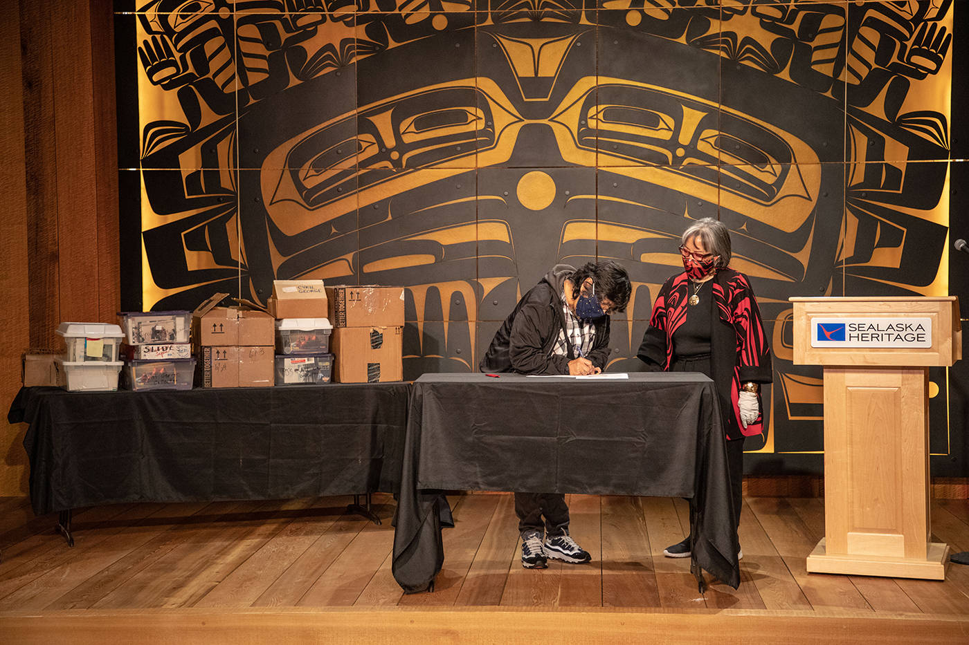 SHI President Rosita Worl and the late Cyril George’s daughter, Roberta Jack, sign a deed of gift at a transfer ceremony last week with the photo collection visible in the background. (Courtesy Photo / Lyndsey Brollini, Sealaska Heritage)