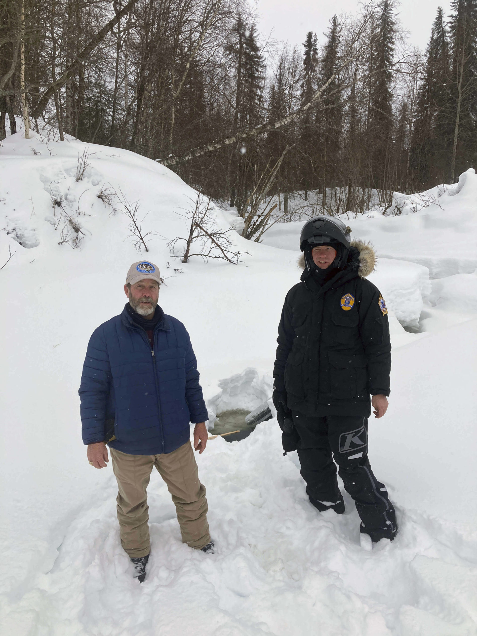 This photo provided by the Alaska Wildlife Troopers taken March 9, 2021, Doug Ramsey, left, of Sundance Wyoming, poses with Alaska Wildlife Trooper Jason Kneier near a hole in the ice of a river in Swentna, Alaska. The two helped pull an 8-year-old boy from the water after he fell into the river. (Alaska Wildlife Troopers)