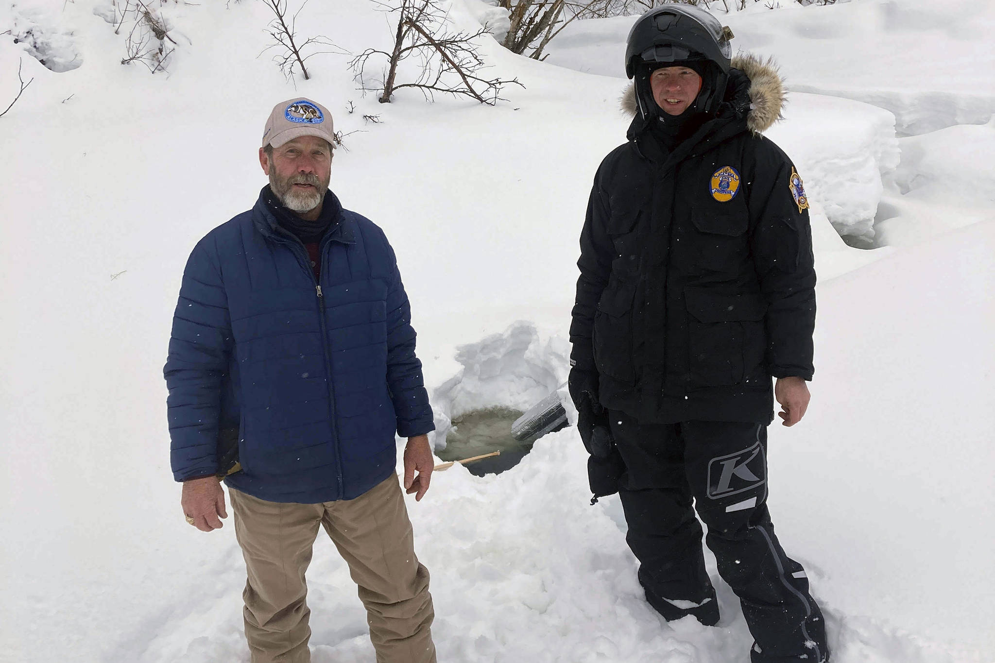 This photo provided by the Alaska Wildlife Troopers taken March 9, 2021, Doug Ramsey, left, of Sundance Wyoming, poses with Alaska Wildlife Trooper Jason Kneier near a hole in the ice of a river in Swentna, Alaska. The two helped pull an 8-year-old boy from the water after he fell into the river. (Alaska Wildlife Troopers)