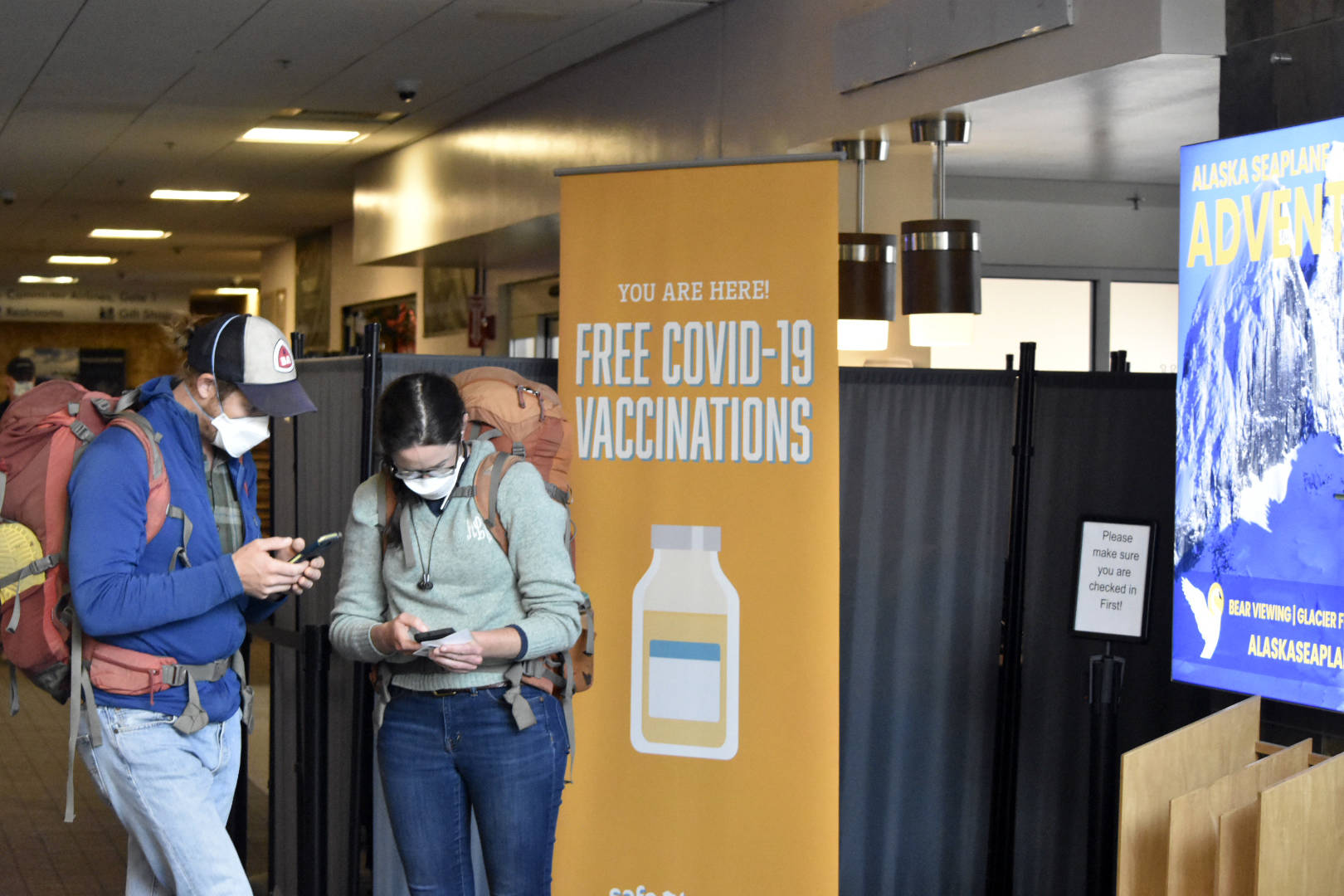 Peter Segall / Juneau Empire 
Travelers check their cellphones near a sign promoting COVID-19 vaccinations in the Juneau International Airport on Thursday. Federal officials are recommending a booster shot for the COVID-19 vaccine, and local health authorities say the rollout will likely be similar to the initial vaccine distribution.