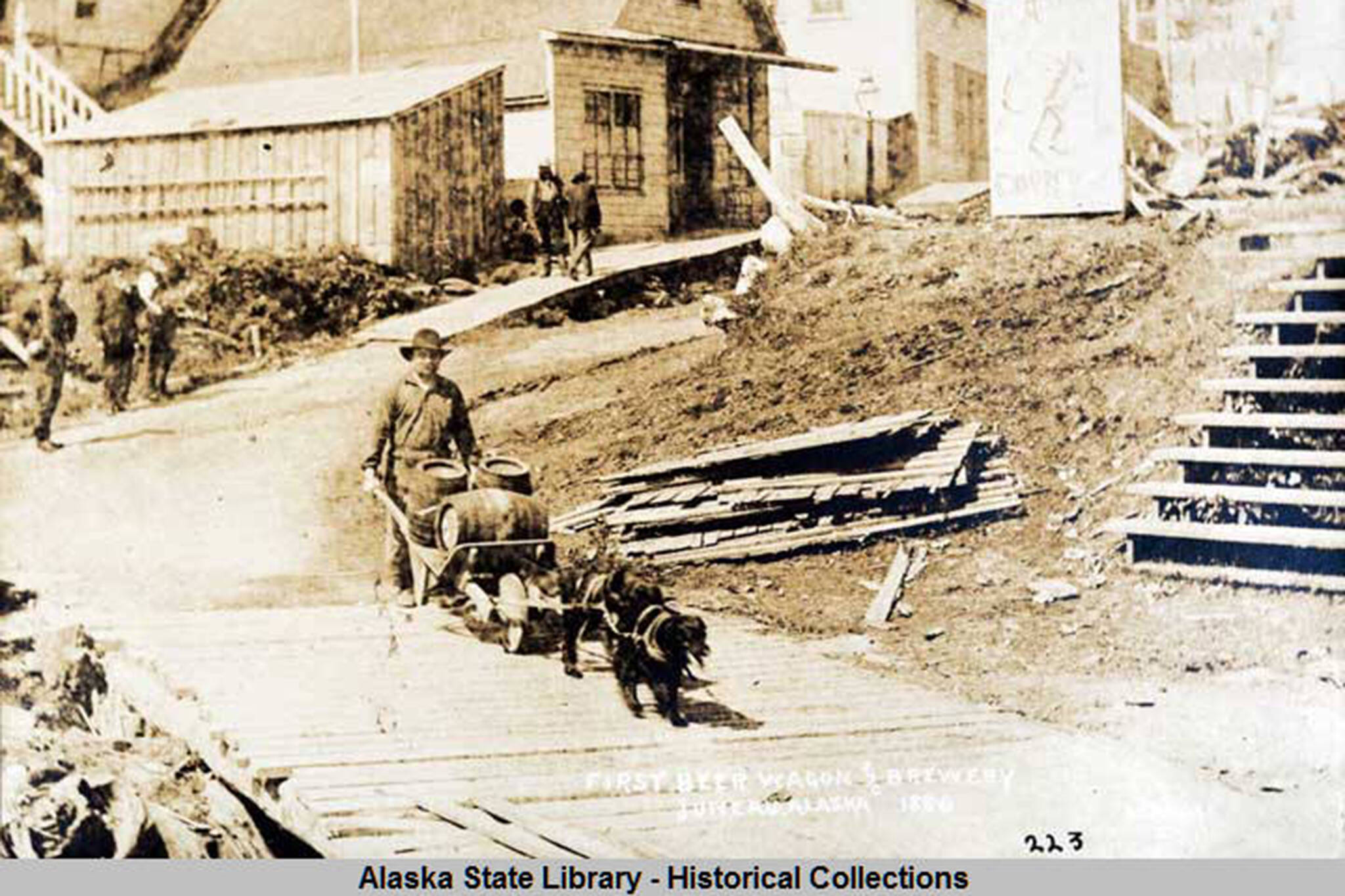 This photo from the Capt. George H. Whitney Photograph Collection shows a man, with wheelbarrow cart and two dogs in harness, transports beer barrels along boardwalk; pedestrians, buildings, and sign for Coon's Drugstore in background in Juneau in 1886. Juneau and Douglas' breweries were the subject of the Gastineau Channel Historical Society's award-winning newsletter. (William Howard Case/ Alaska State Library - Historical Collections)