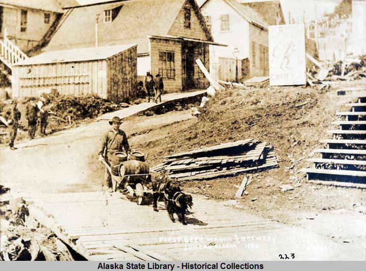 This photo from the Capt. George H. Whitney Photograph Collection shows a man, with wheelbarrow cart and two dogs in harness, transports beer barrels along boardwalk; pedestrians, buildings, and sign for Coon’s Drugstore in background in Juneau in 1886. Juneau and Douglas’ breweries were the subject of the Gastineau Channel Historical Society’s award-winning newsletter. (William Howard Case/ Alaska State Library - Historical Collections)