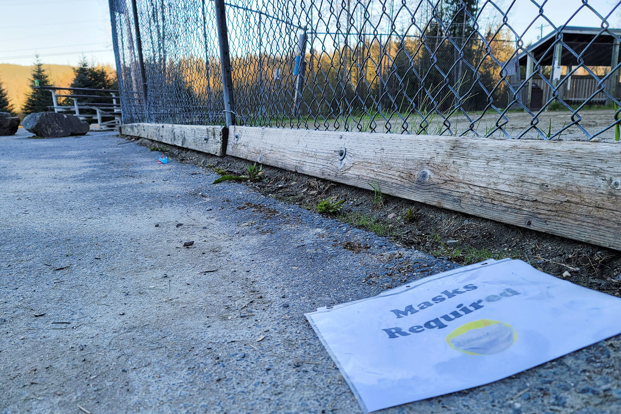 A sign reading “Masks Required” lies on the ground near a softball field at Melvin Park in this May photo. The City and Borough of Juneau is lowering the community risk level for COVID-19, but masks remain required inside public spaces. (Ben Hohenstatt / Juneau Empire)