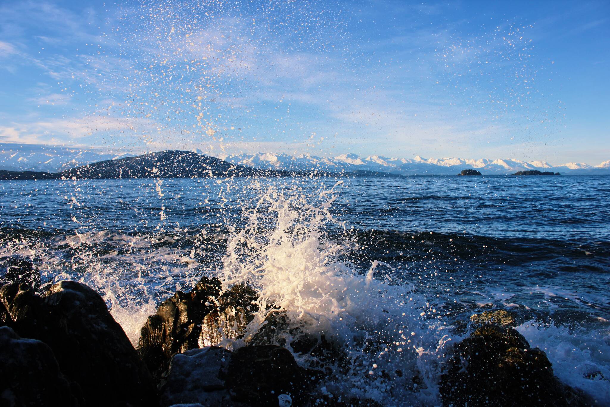 A wave breaks over the rocky shores of Lynn Canal, with Shelter Island and the Chilkat Range seen on the horizon in Southeast Alaska on Dec. 13, 2020. (Courtesy Photo / Sally Thompson)