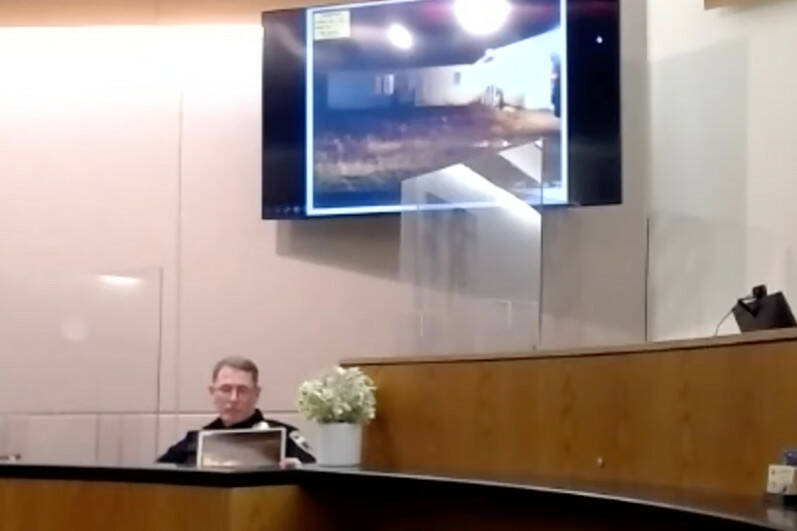 Officer John Waldron of the Yakutat Borough Police Department looks at a print of a photograph as he testifies about a stabbing he responded to in 2018. (Screenshot)