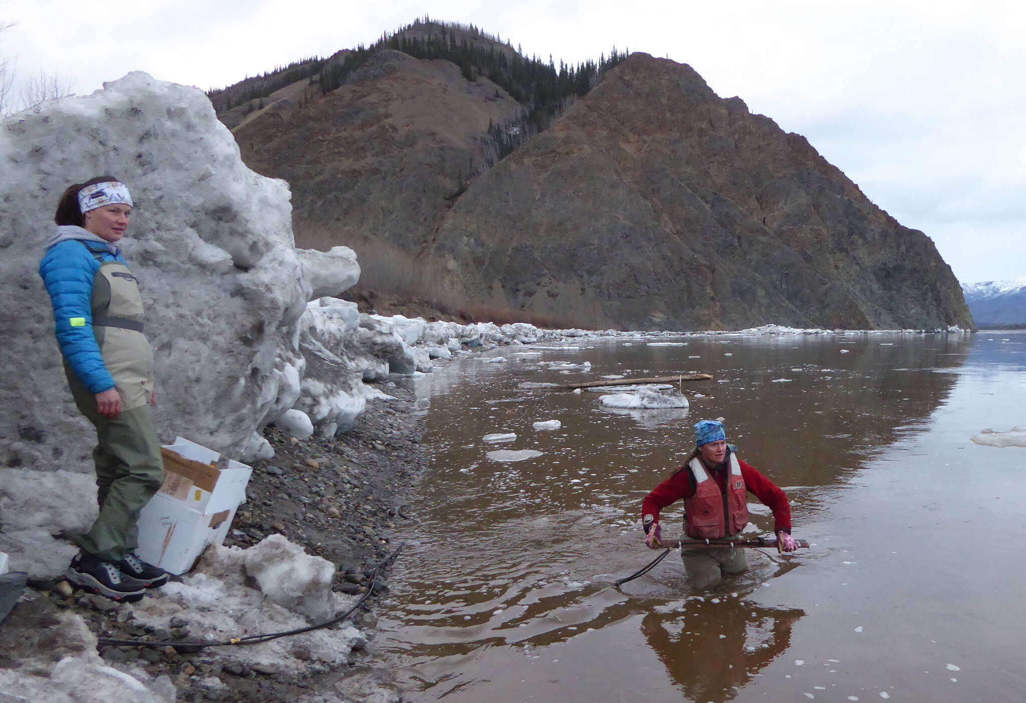Heather Best (in water), a USGS hydrologist, prepares to toss a road-grader blade with a river-measuring device attached into the Yukon River near Eagle, Alaska. USGS hydrologic technician Liz Richards watches for icebergs. (Courtesy Photo / Ned Rozell)
