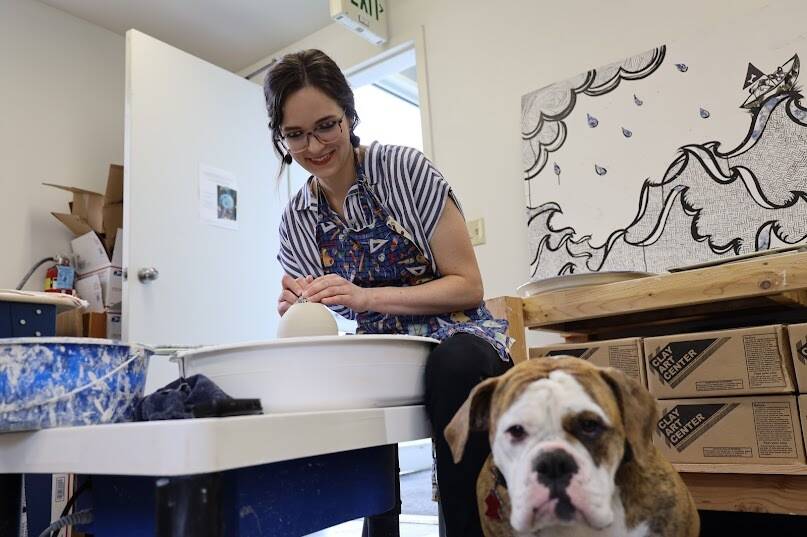 Mercedes Muñoz smiles at her dog, Coco, as she works on shaping ceramic pieces on her potter’s wheel. Muñoz is set to host first ever show at the her late grandmother’s galley, the Rie Muñoz Gallery, on Sunday which will feature her summer collection of work. (Clarise Larson / Juneau Empire)