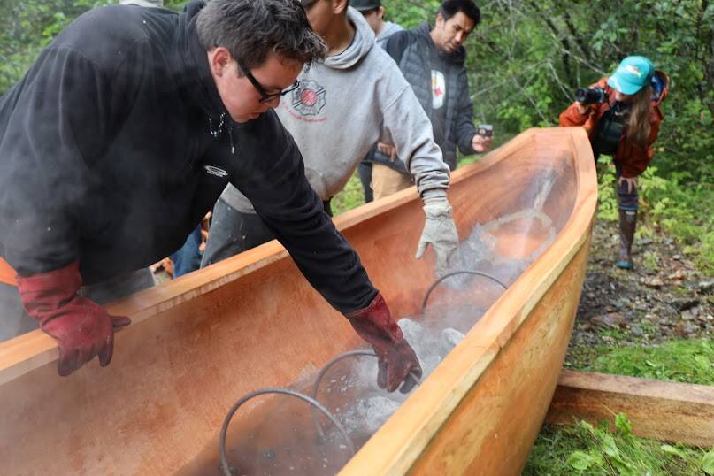 Anthony Johnson, a senior at Chatham School District, reaches down to grab the metal cages that keeps the rocks from burning through the wood. This is Johnson’s second time experiencing the steaming of a canoe as he also participated in a dugout steaming in Hoonah in 2016. (Clarise Larson / Juneau Empire)
