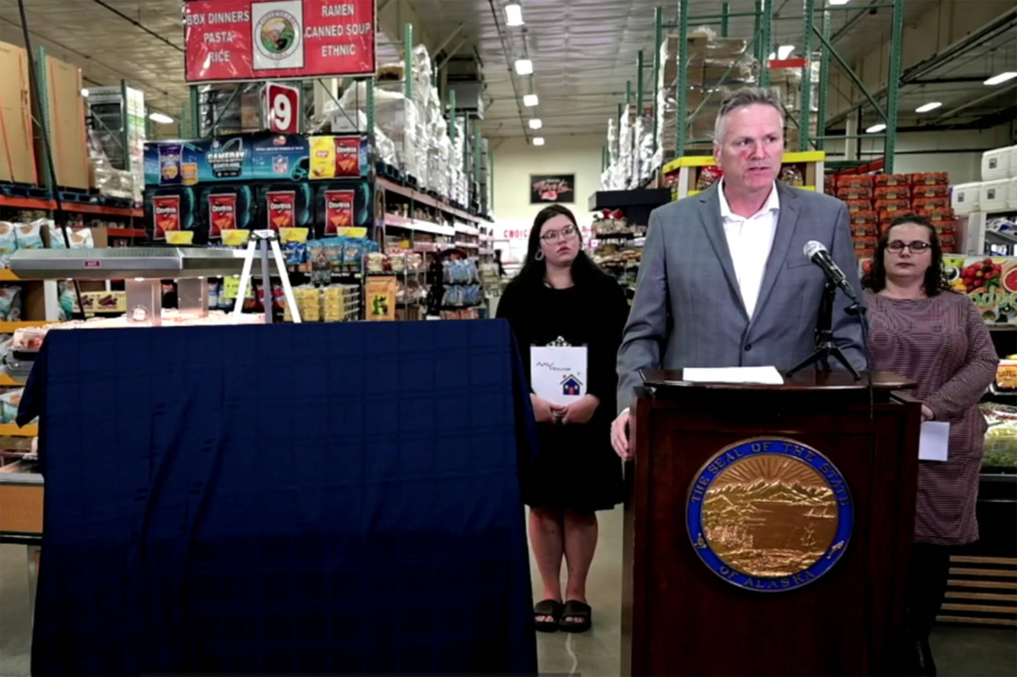 Gov. Mike Dunleavy stands at a lectern in a Three Bears Alaska store in Palmer, next to a giant, covered check displaying this year’s Permanent Fund dividend amount. Behind him stand Alaska resident Miranda Wagoner (left) and Jessica Viera, executive director, Wasilla Chamber of Commerce (right) who gave speeches at the event. (Faceboook live screenshot)