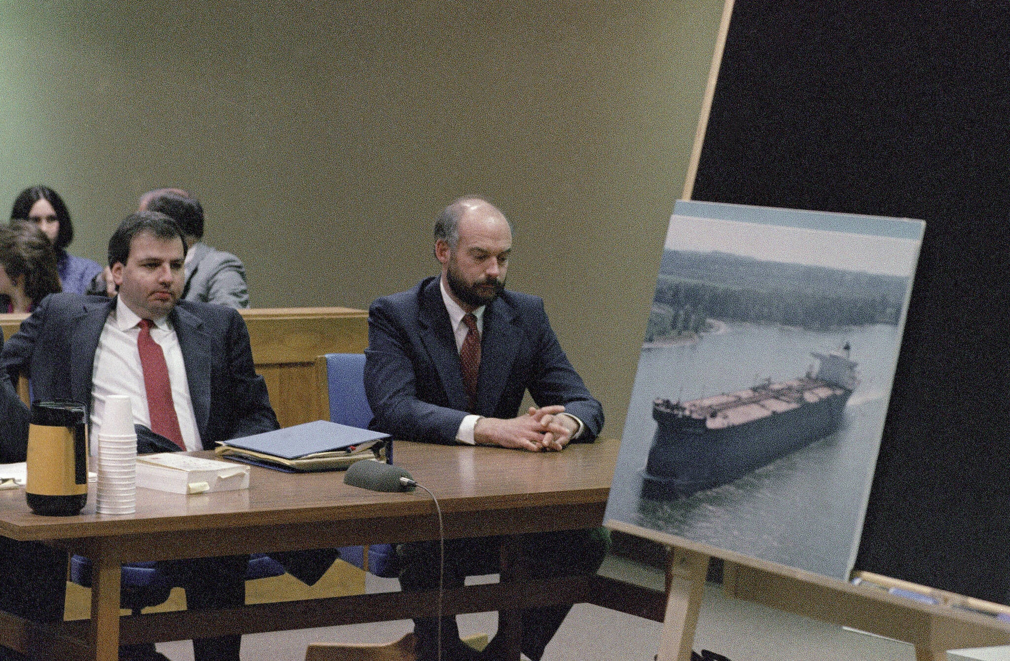 Fired Exxon Valdez skipper Joseph Hazelwood, right, sits with his lawyer Michael Chalos as a photograph of the tanker is displayed on an easel in Anchorage Superior Court, March 20, 1990. Hazelwood, the captain of the Exxon Valdez oil tanker that ran aground more than three decades ago in Alaska, causing one of the worst oil spills in U.S. history, has died in July 2022, the New York Times reported. He was 75. (AP File Photo / Jack Smith)