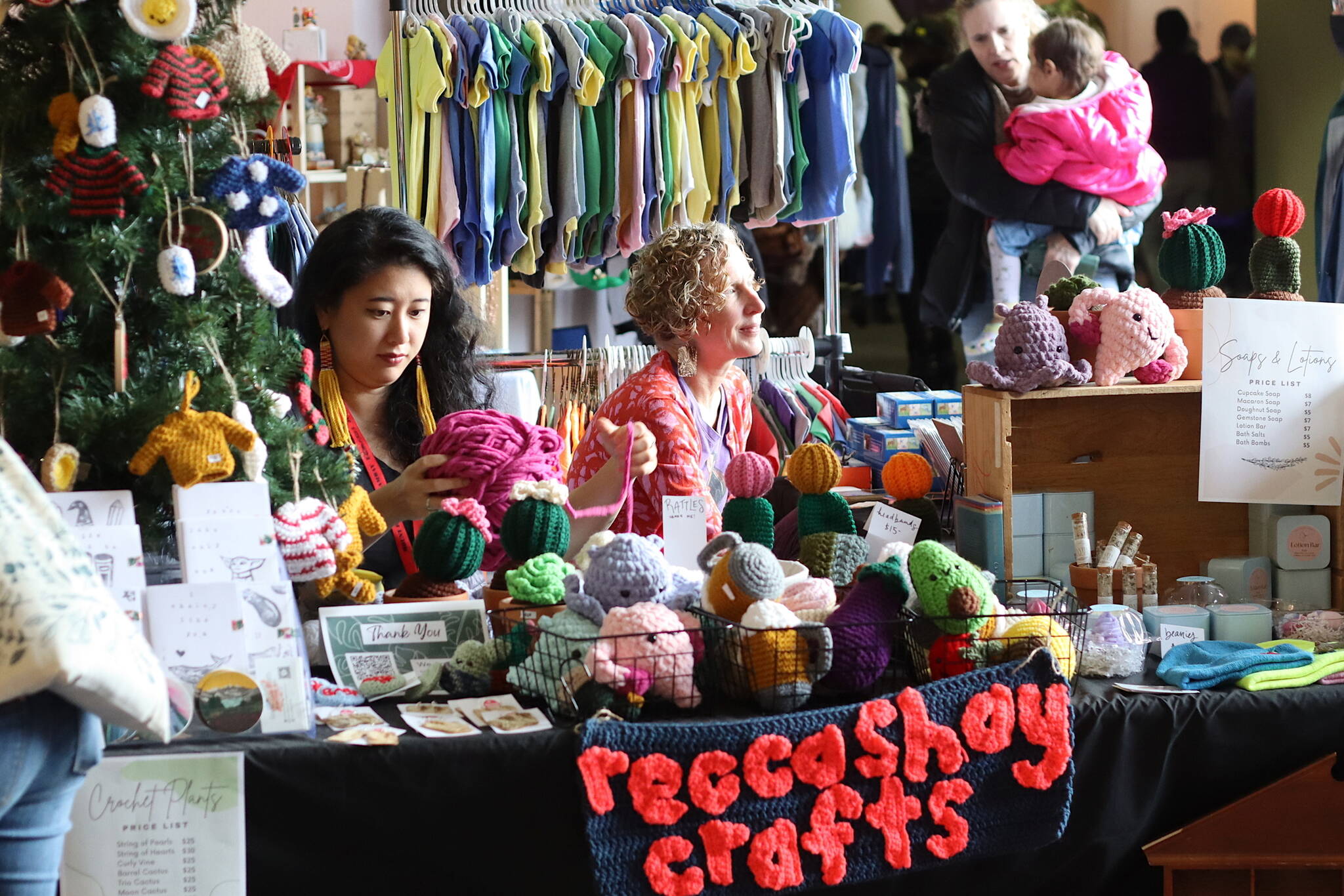 Rebecca Hsieh, left, knits small gifts as a first-time vendor at the Juneau Public Market as her friend, MK MacNaughton, a longtime vendor sells hand-painted items near the entrance of Centennial Hall on Saturday. The market, celebrating its 40th year, is scheduled to continue from 10 a.m. to 5 p.m. Sunday. (Mark Sabbatini / Juneau Empire)