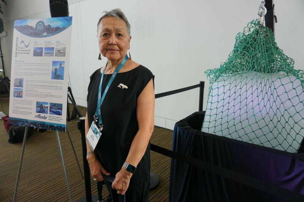Vera Metcalf stands on Wednesday by a chunk of sea ice transported from Utqiagvik and displayed at the Arctic Encounter Symposium. The melting ice, which started at 310 pounds, symbolizes the rapid climate change that is weaking the Arctic ice pack, with profound implications for ecosystems, communties and cultures. (Photo by Yereth Rosen / Alaska Beacon)