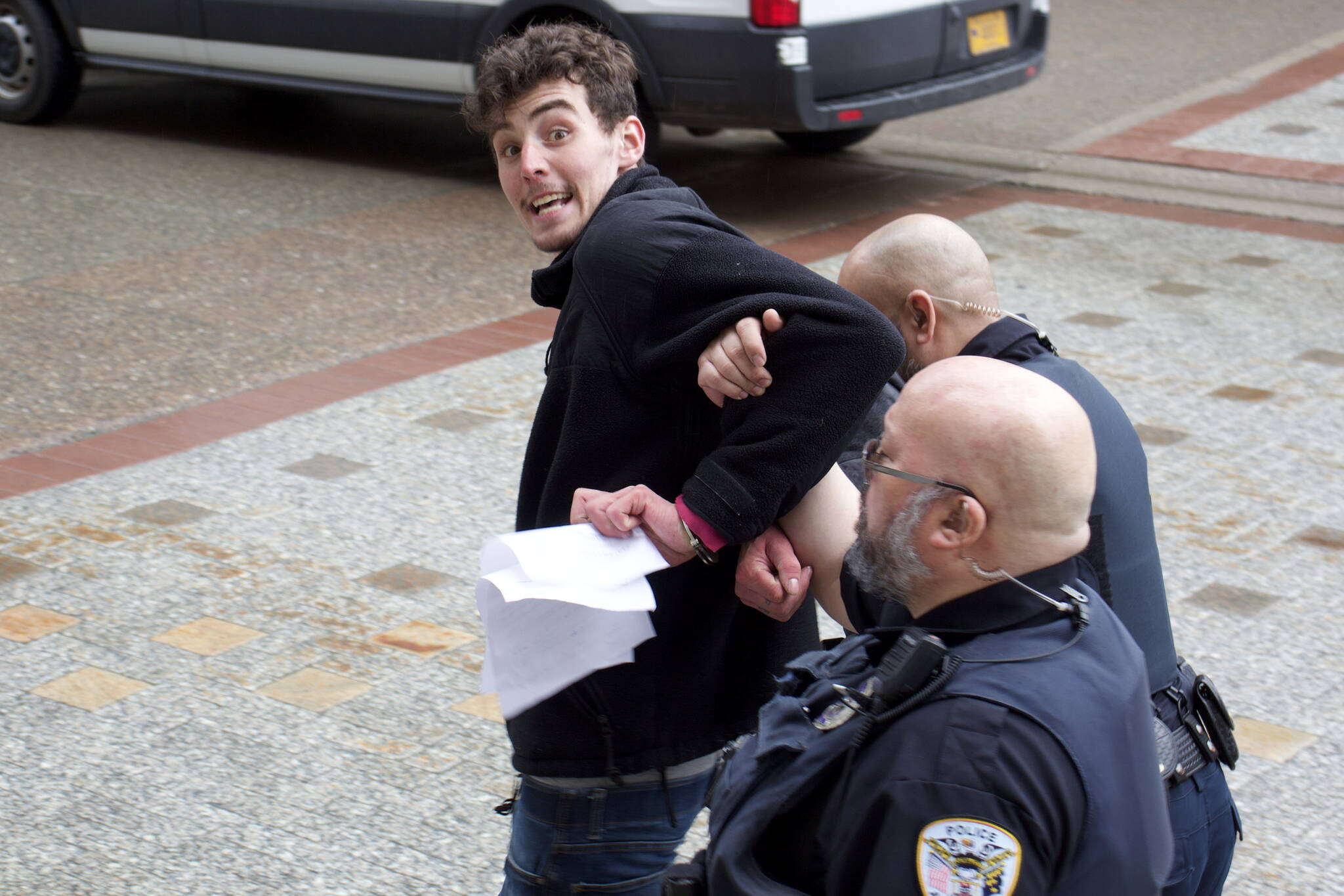 Eric Osuch tries to offer papers related to his arrest in front of the Alaska State Capitol on Monday morning to a reporter as Juneau Police Department officers escort him to a nearby patrol vehicle. Osuch, who was staging a solo protest about fisheries bycatch policies, was banned from the Capitol after causing a public disruption and was arrested a short time later for another alleged disturbance inside the State Office Building. (Mark Sabbatini / Juneau Empire)