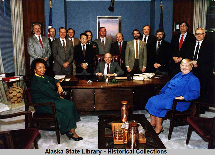 This photo held by the Alaska State Library shows the cabinet of Gov. Bill Sheffield. Seated in front of desk, left – Eleanor Andrews (Administration), right - Esther Wunnicke (Natural Resources). Standing – Marshall Lind (Education), Gary Hayden (Marine Highway System), Jim Robison (Labor), Roger Endell (Corrections), Emil Notti (Community & Regional Affairs), Gen. Edward Pagano (Adjutant Gen., Miltary & Veterans Affairs), Loren H. Lounsbury (Commerce & Econ. Dev.), Richard Knapp (DOTPF), Bill Ross (DEC), Don Collinsworth (F&G), John Pugh (H&SS), Hal Brown (Attorney General, Law), Robert Sundberg (Public Safety).  (Alaska Office of the Governor Photograph Collection, 1959 to present. ASL-PCA-213)