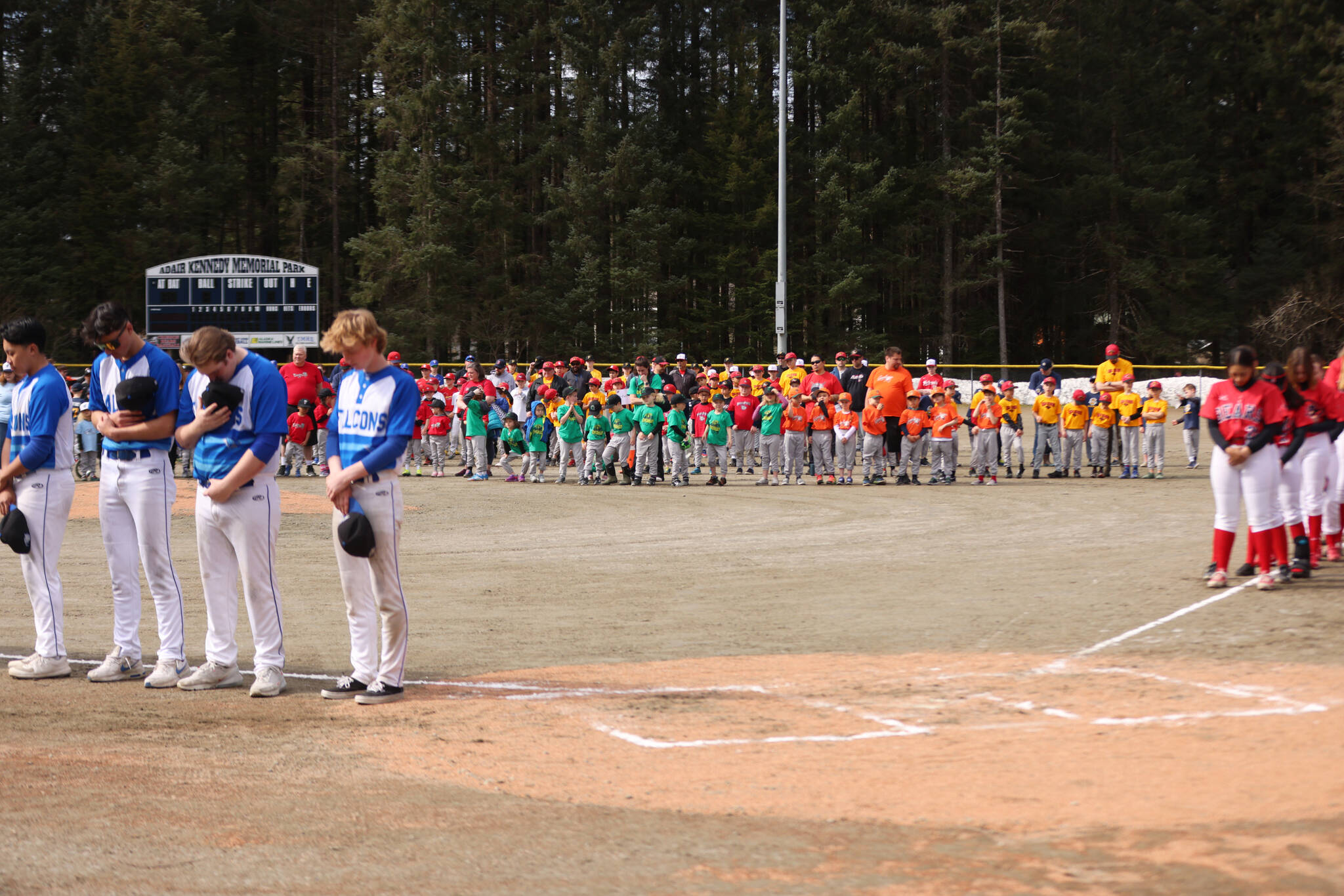 High school and Little League baseball and softball teams bow their heads during the invocation that was part of Gastineau Channel Little League’s opening day Saturday at Adair-Kennedy Memorial Park. Invocation was given by Charles Rohrbacher, deacon for the Cathedral of the Nativity of the Blessed Virgin Mary. This year, nearly 450 youths are participating in GCLL. (Ben Hohenstatt / Juneau Empire)