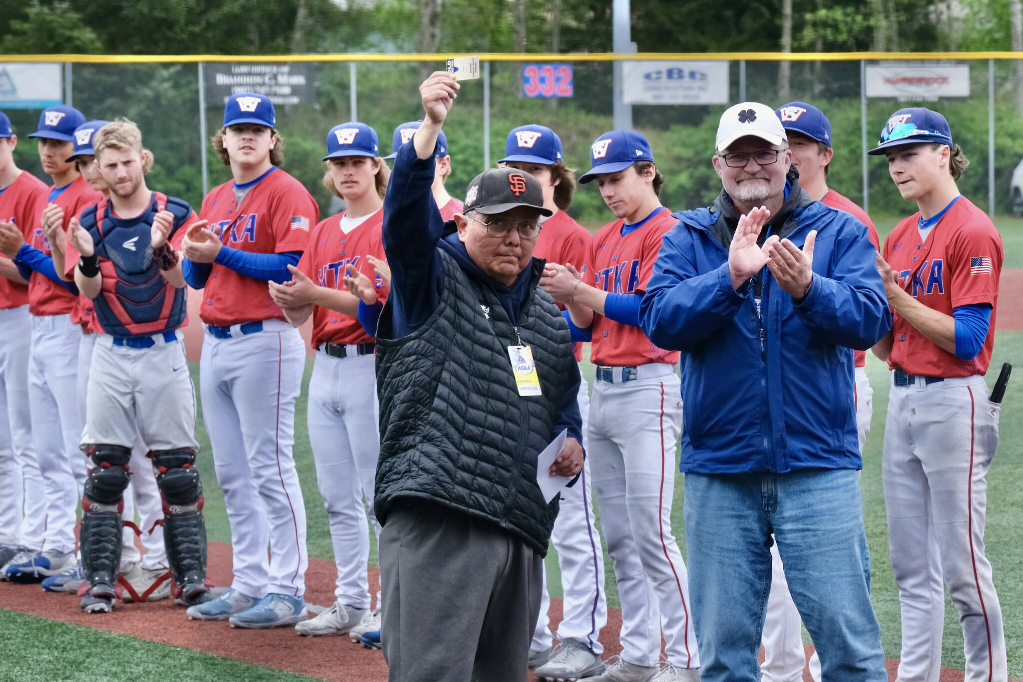 Lawrence “Woody” Widmark holds up his Alaska School Activities Association Gold Lifetime Pass as Sitka Wolves players and ASAA executive director Billy Strickland applaud during the presentation at the ASAA State Baseball Championships in Sitka last week. (Klas Stolpe / Juneau Empire)