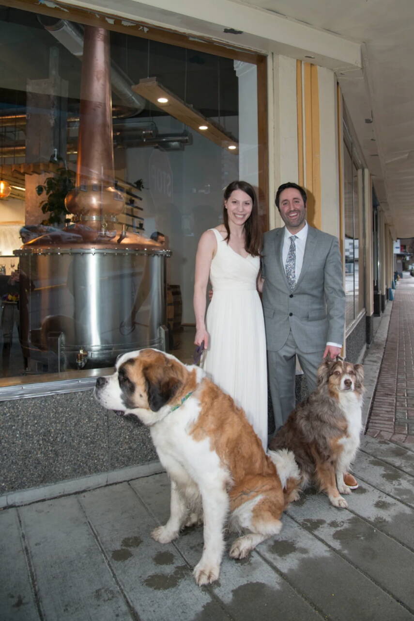 Maura Selenak and Brandon Howard celebrate their marriage in front of the Amalga Distillery they have created together in the historic 1937 AELP building. They stand — with their dogs Walter and Wallaby — on their wedding day in 2017 on the corner of Second and Seward Streets with the custom-built pot still, made in Kentucky, visible through the large front window. (Photo by Ben Huff, courtesy of Maura Selenak)