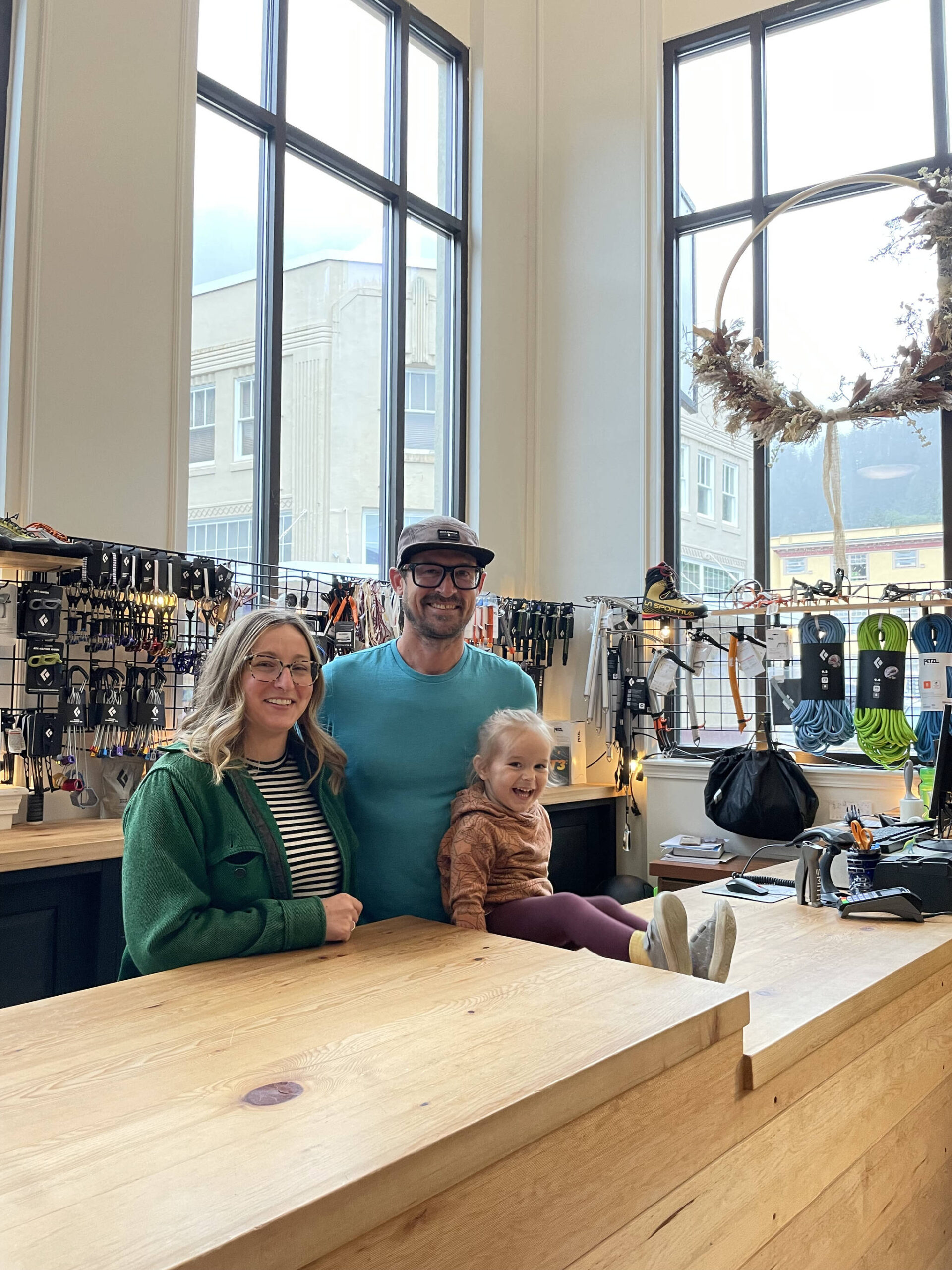 Foggy Mountain Shop owners Sean Reilly and Courtney Nicholl with their daughter Alder stand at their custom-built Icy Straits Lumber counter. Courtney designed the space to focus attention on inspiring outdoors activities with the steep mountain backdrop. (Photo by Laurie Craig)