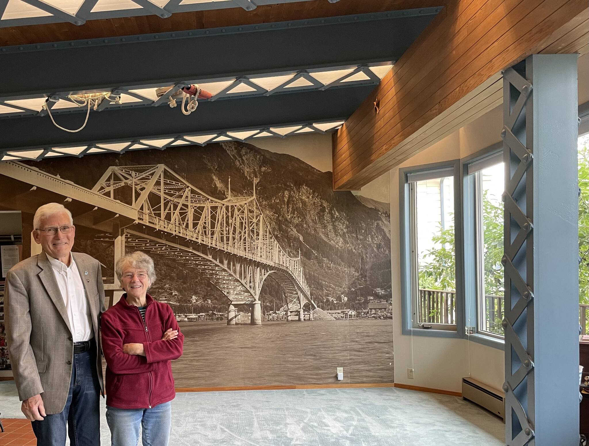 Homeowners Bill Leighty and Nancy Waterman stand in their living room in September of 2023. A mural of an enlarged Winter and Pond photo of the original Douglas Bridge provides background for structural elements from the bridge used in their home built in 1983. (Photo by Laurie Craig)