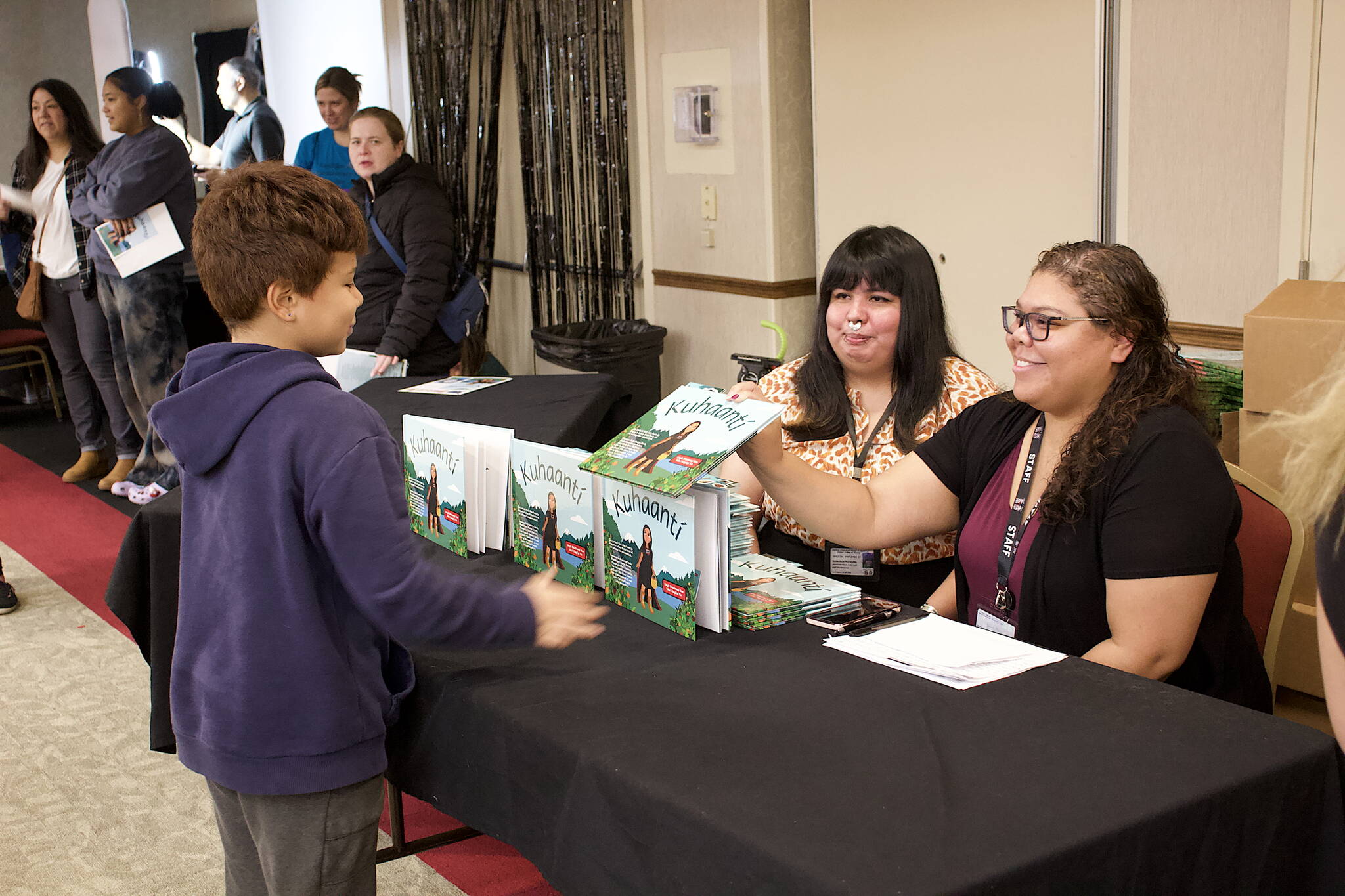 Felicia Price, an employee of the Central Council of Tlingit and Haida Indian Tribes of Alaska, hands a copy of the Lingít-language book “Kuhaantí” to her son, Brayden, 8, while staffing the distribution table for the book with co-worker Genevieve McFadden during its release party Friday night at Elizabeth Peratrovich Hall. (Mark Sabbatini / Juneau Empire)